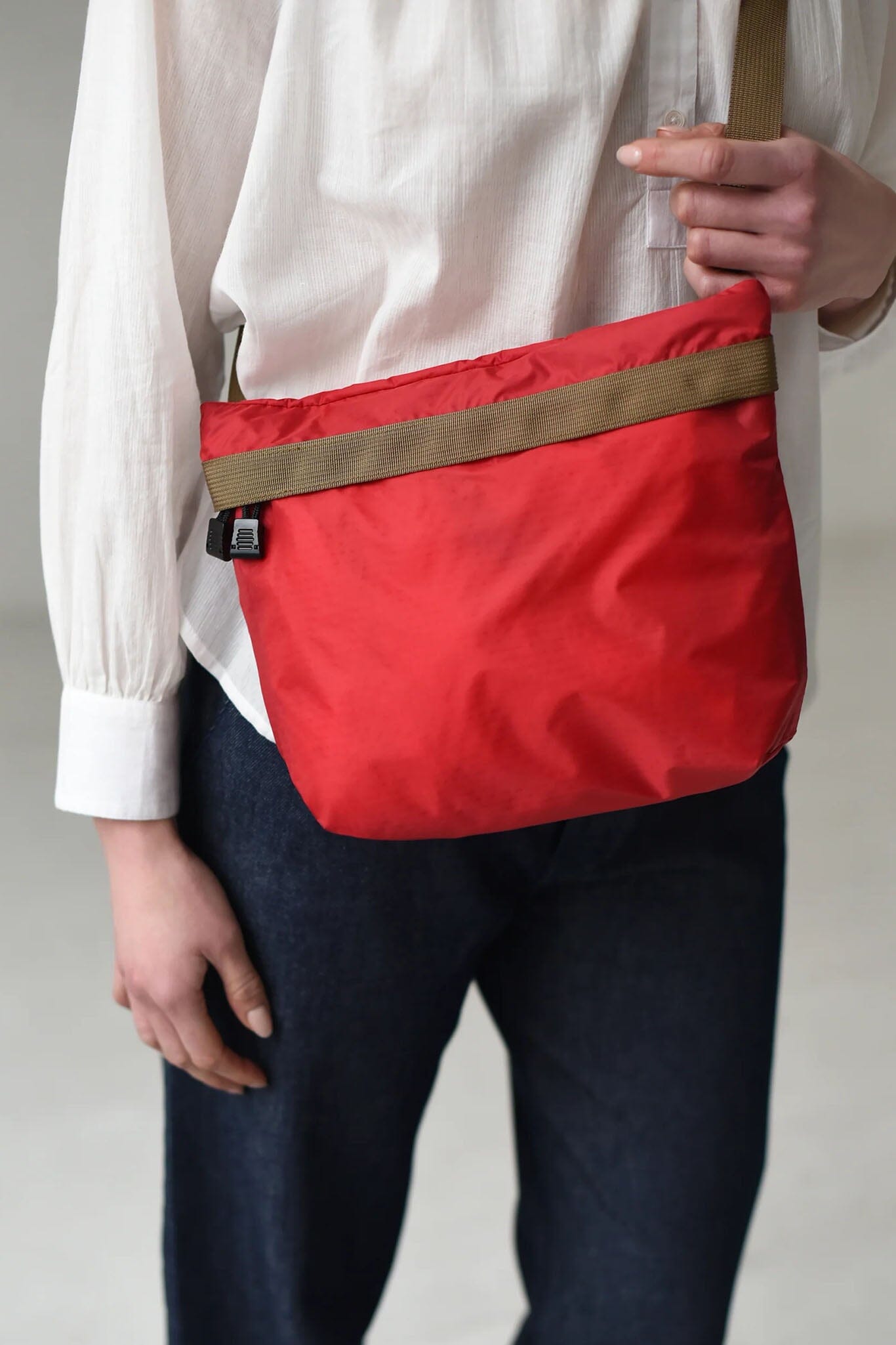Red bag with a brown strap held by a person wearing a white shirt and dark pants on a plain background