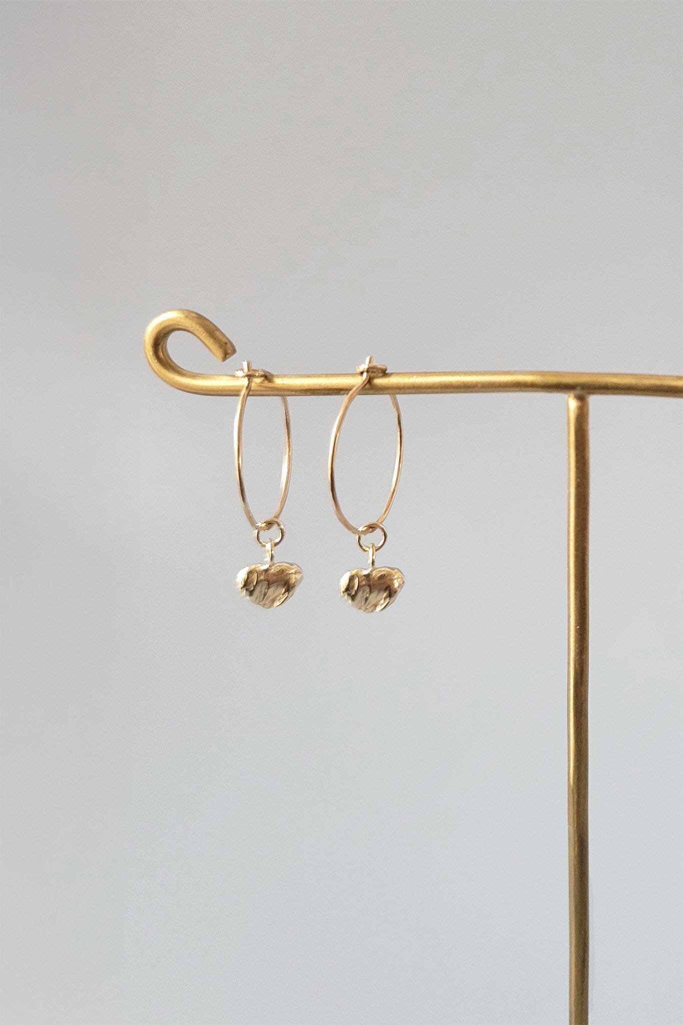 Gold hoop earrings with heart-shaped charms on a gold stand against a gray background