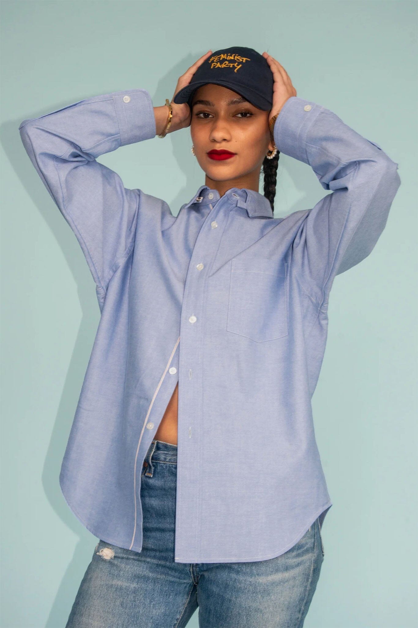 Person wearing a light blue button-up shirt and jeans with a black cap on a light blue background
