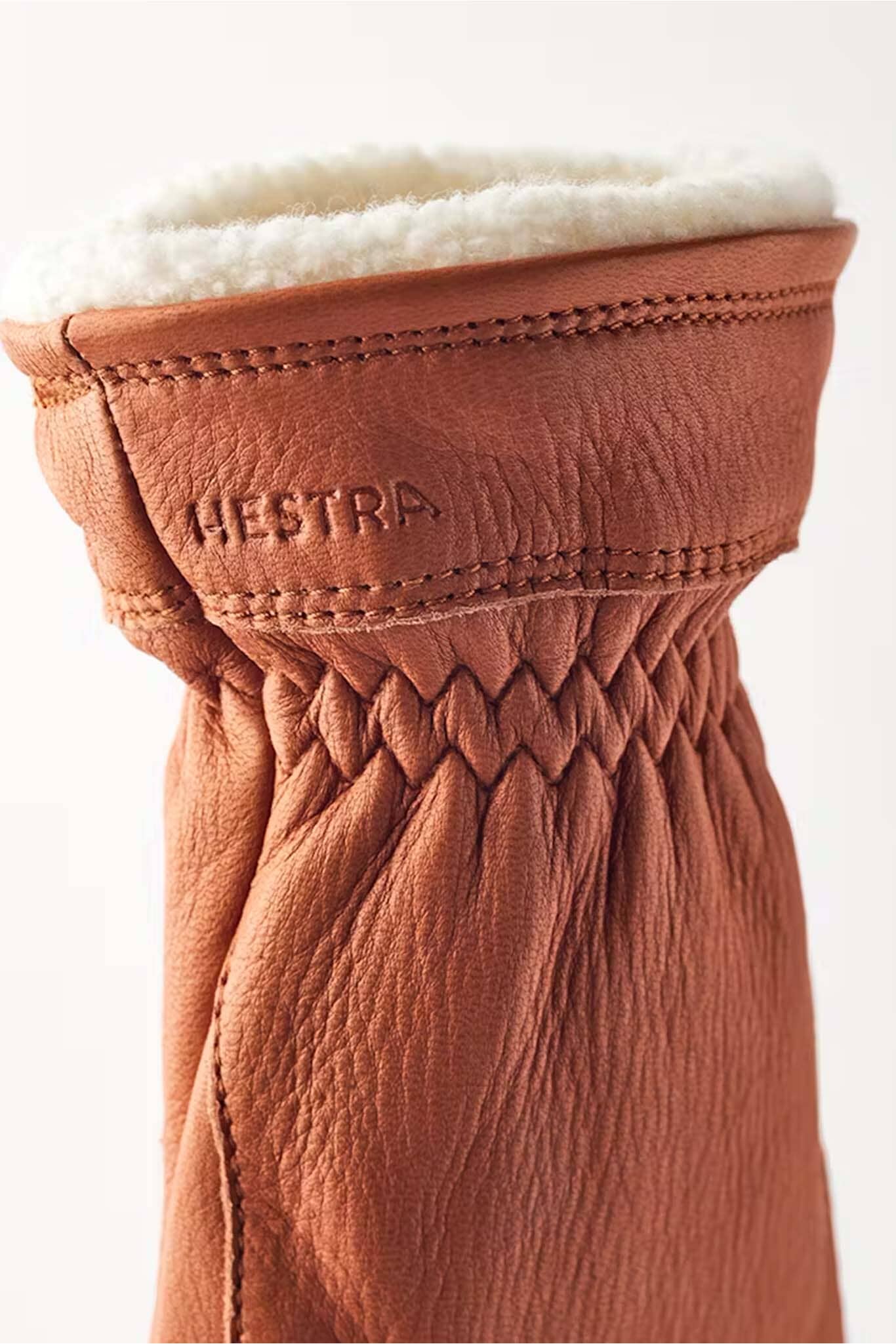Close-up of a brown leather glove with white fur lining, featuring the Hestra brand logo.