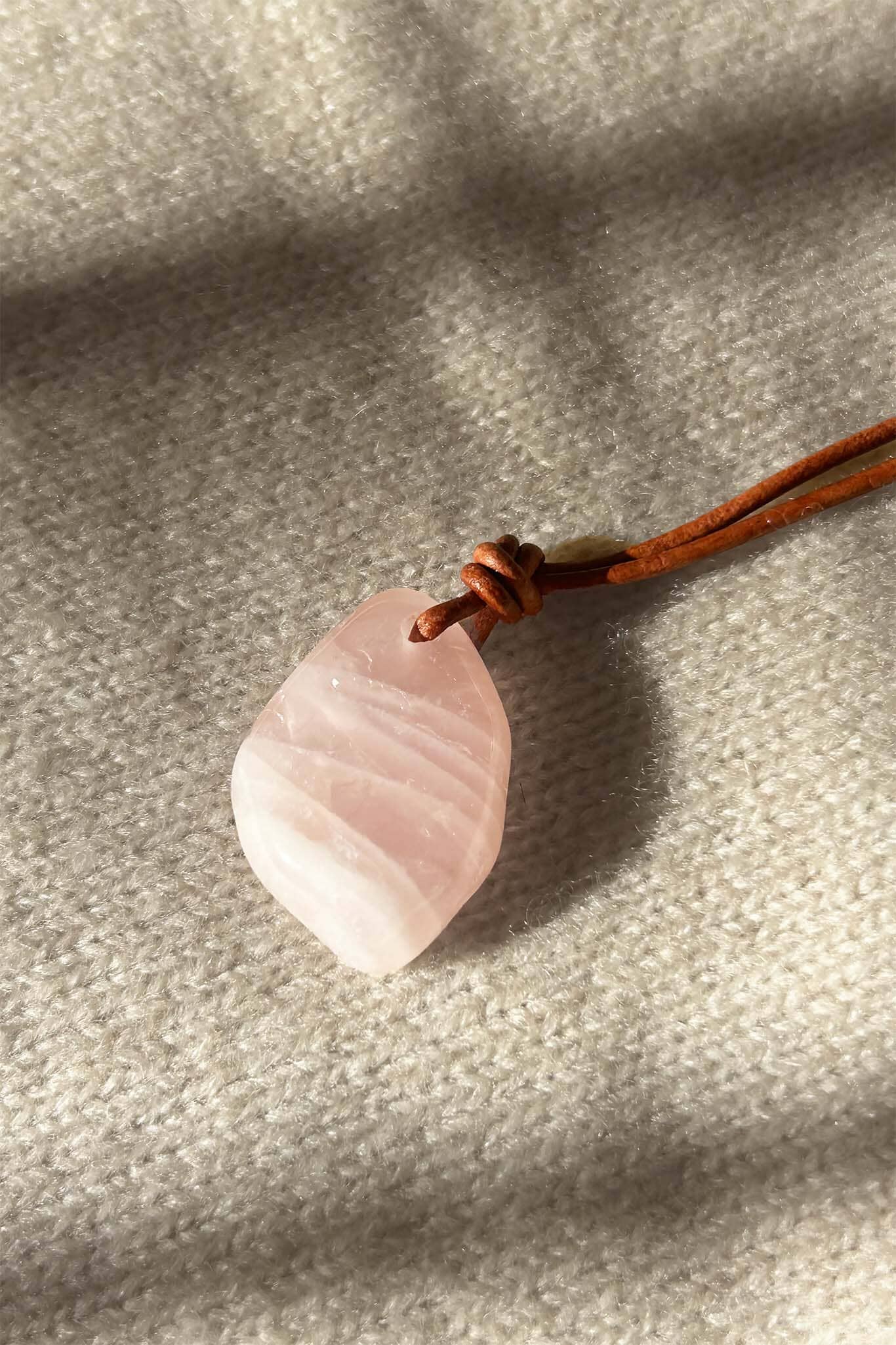 Pink rose quartz stone pendant on a brown cord against a gray fabric background
