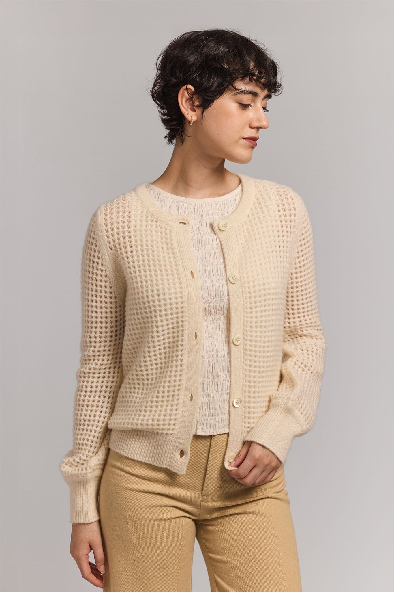 Woman wearing a cream textured cardigan over a white shirt with beige pants on a gray background