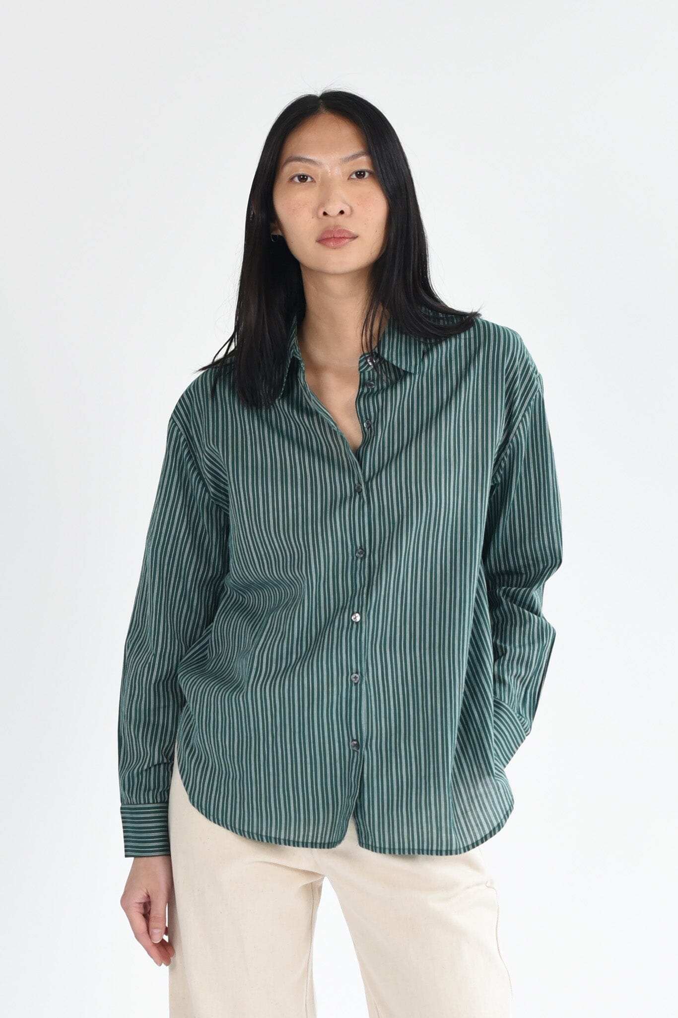 A woman wears a dark green, striped long sleeve button down blouse. The top is lightweight and made of cotton. Available to purchase at M.Patmos in Brooklyn, New York.
