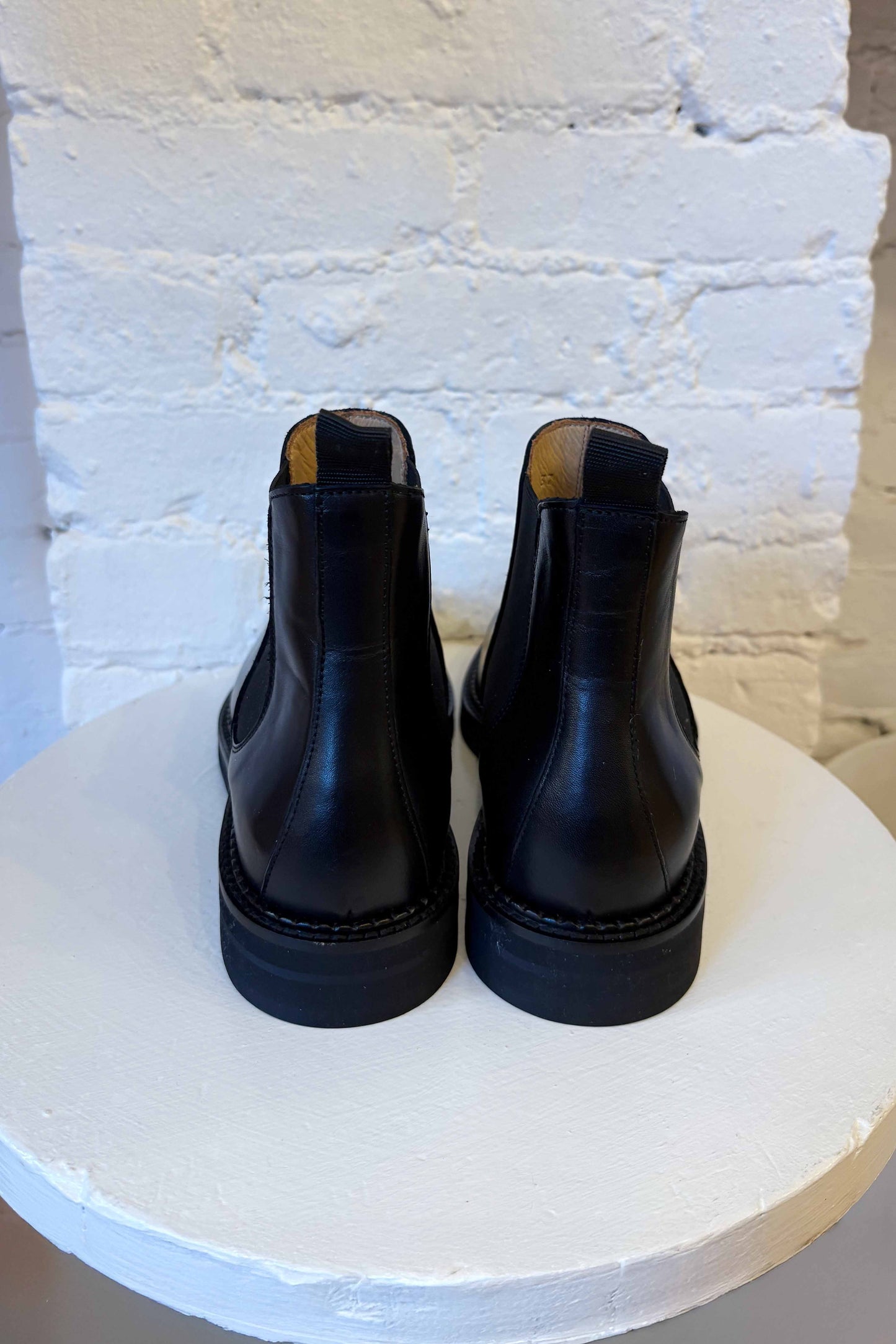 Pair of black leather boots on a white pedestal against a light blue brick wall.