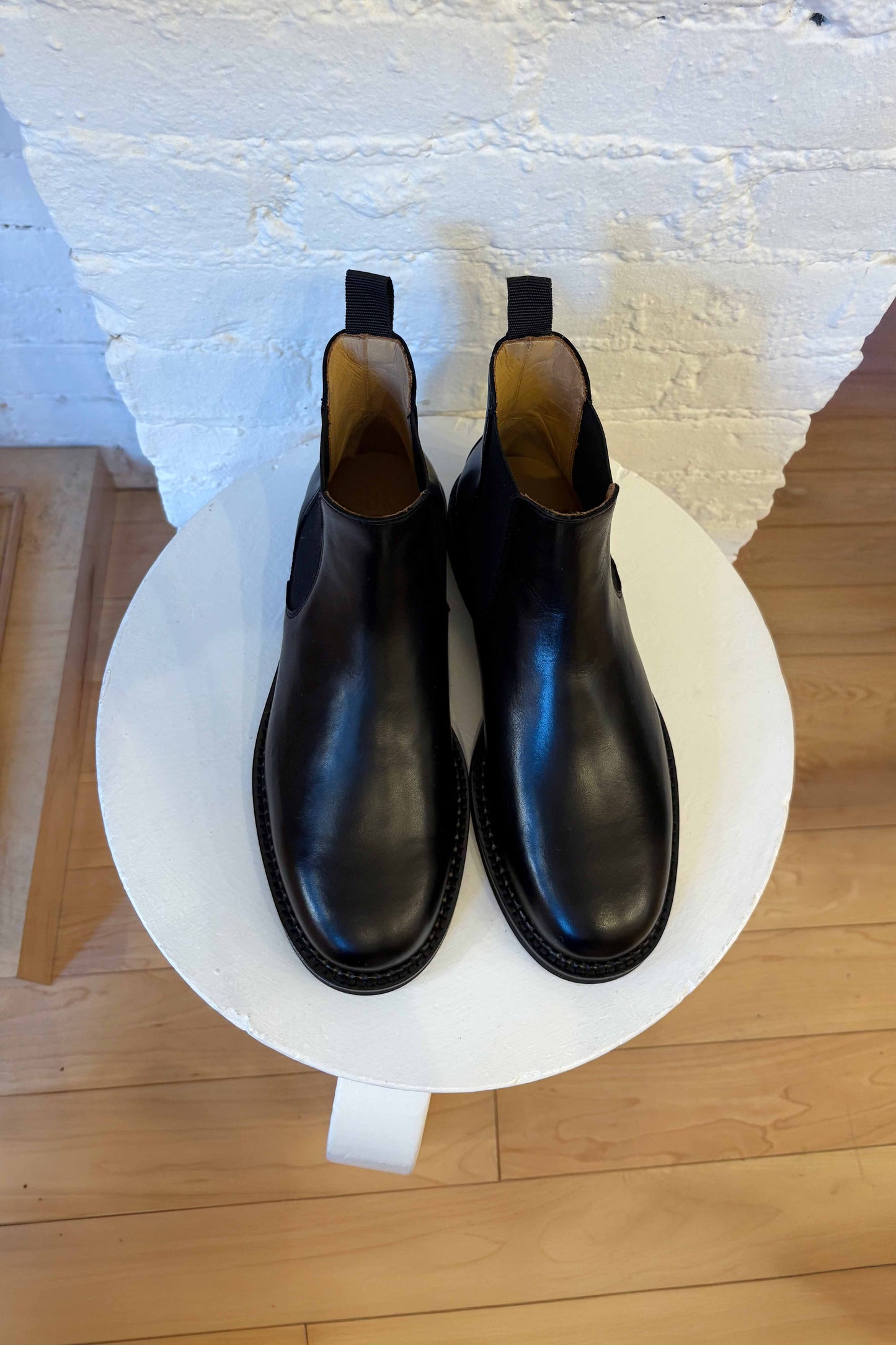 Pair of black leather boots on a white circular platform with a light wood floor and white wall background.