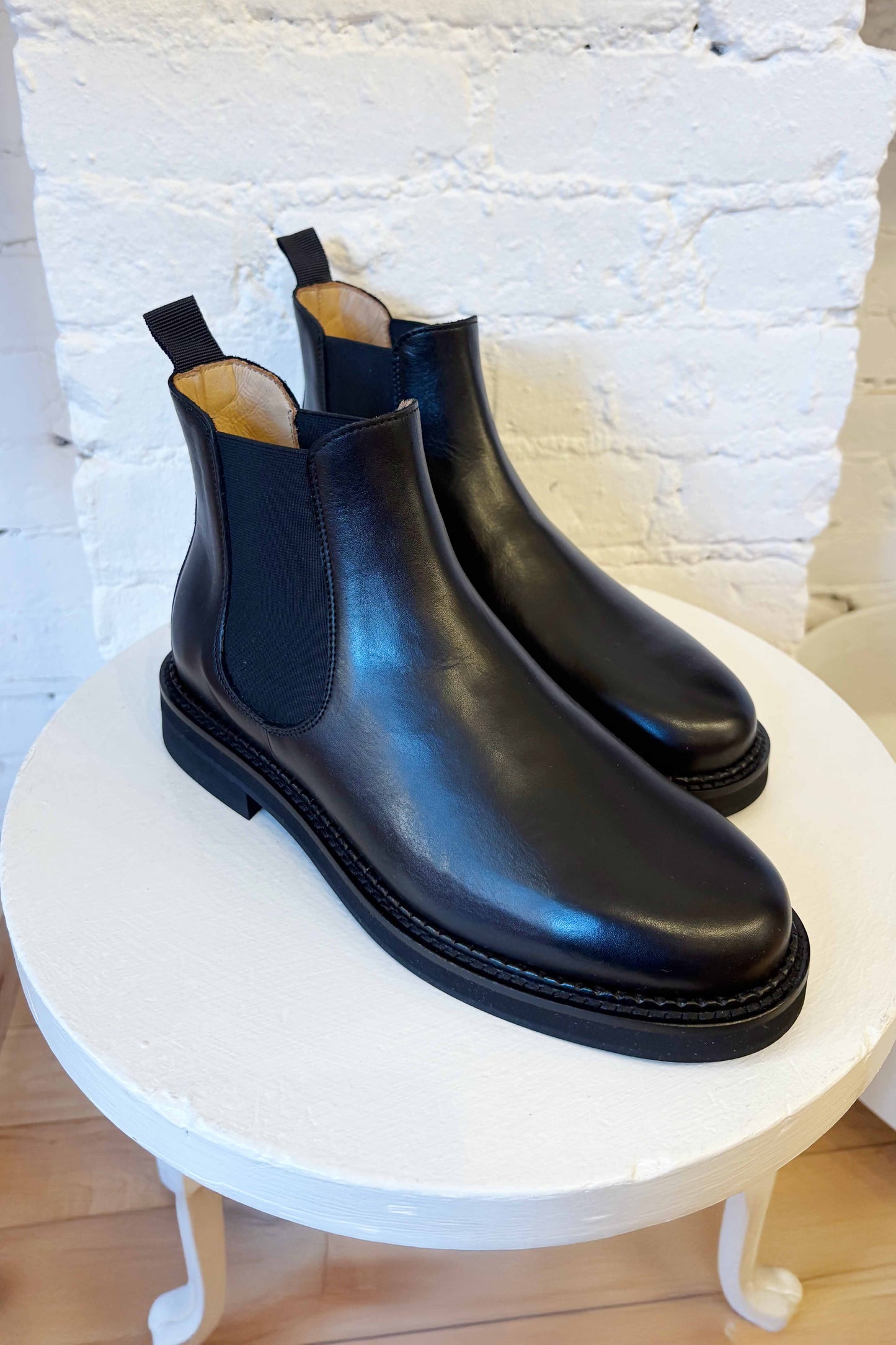 Pair of black leather Chelsea boots on a small round table with a white brick wall background.