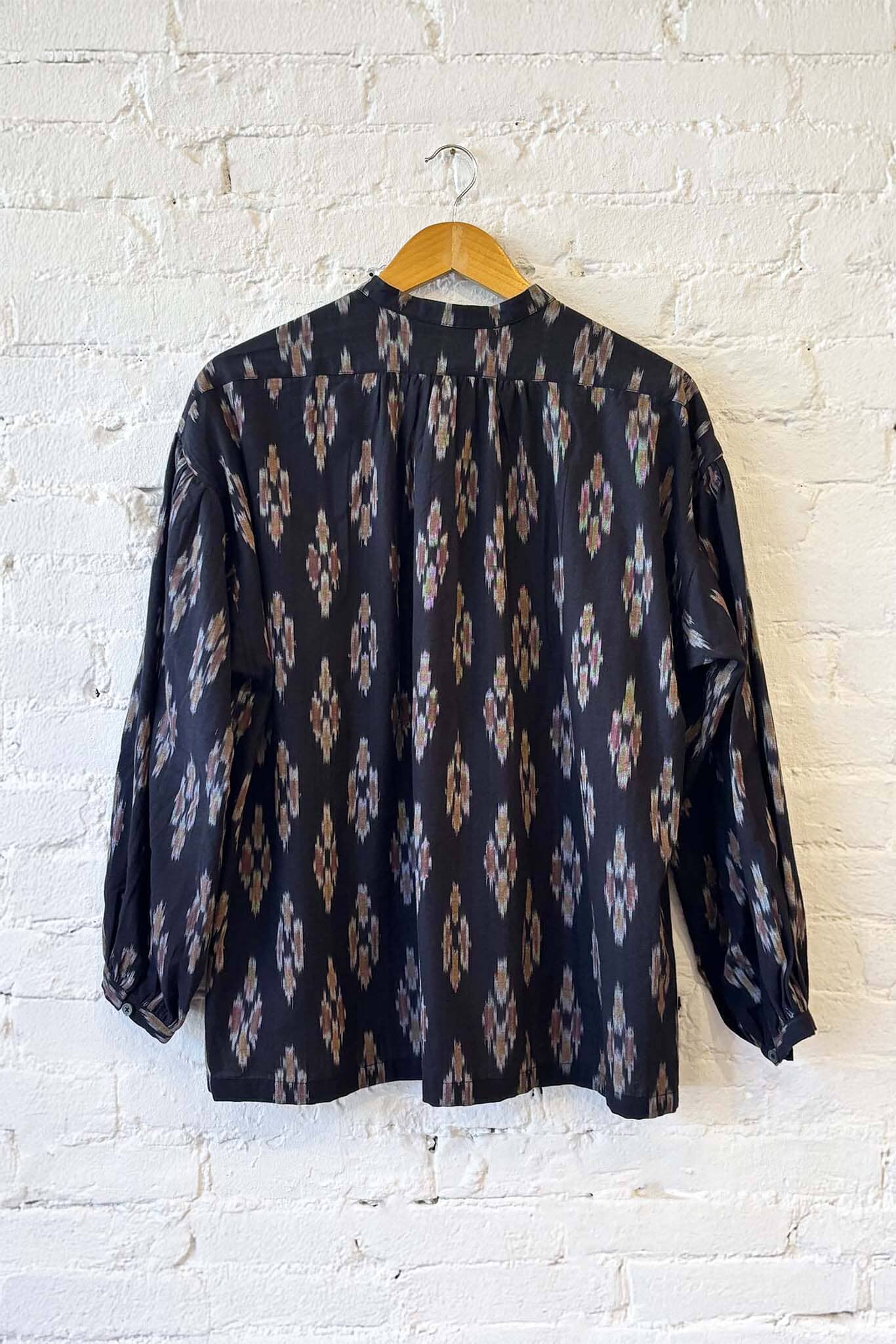 The back view of a black patterned blouse on a wooden hanger against a white brick wall.