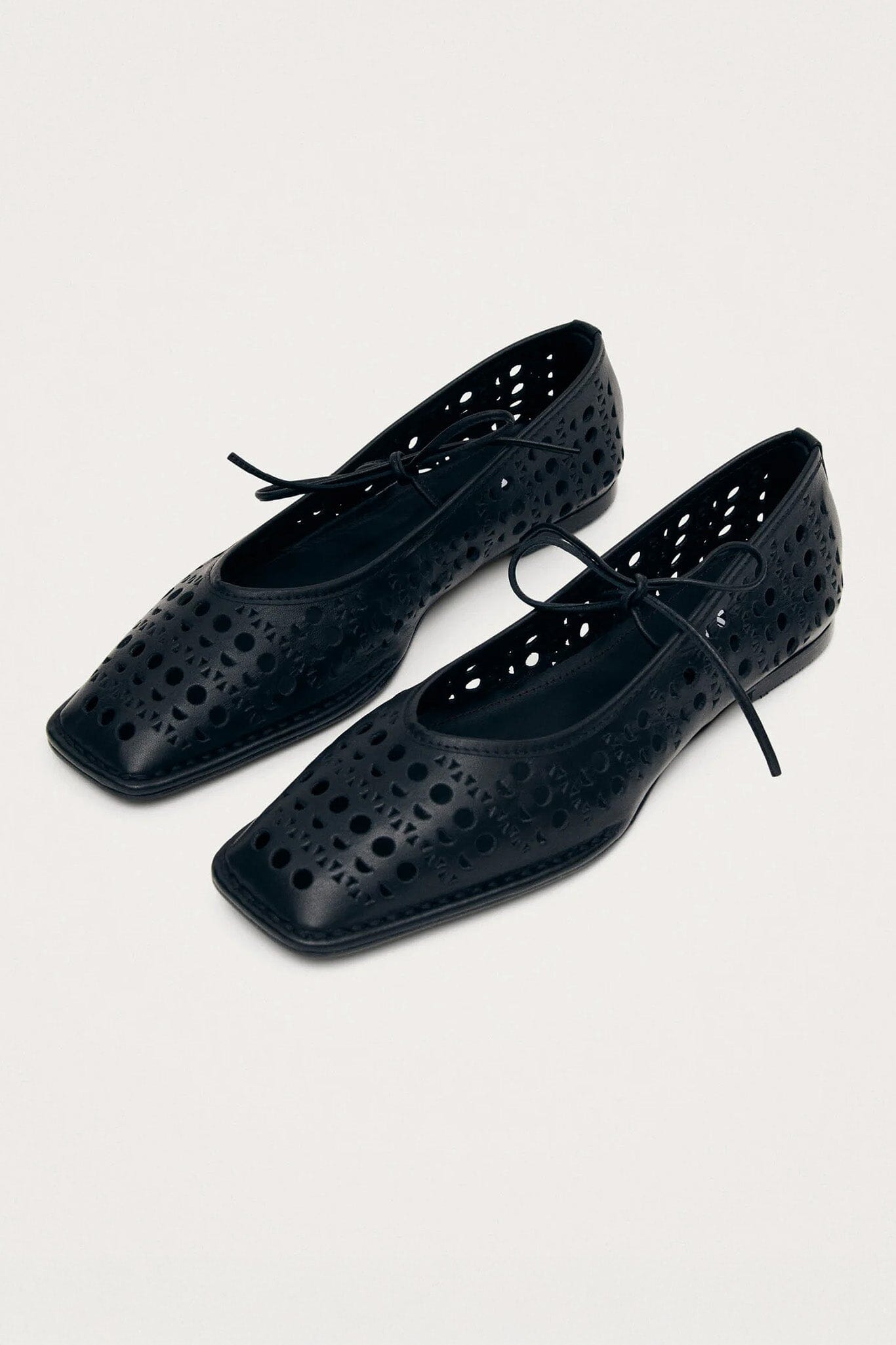 A pair of black leather ballet flats with laser-cut patterns and a bow detail on top