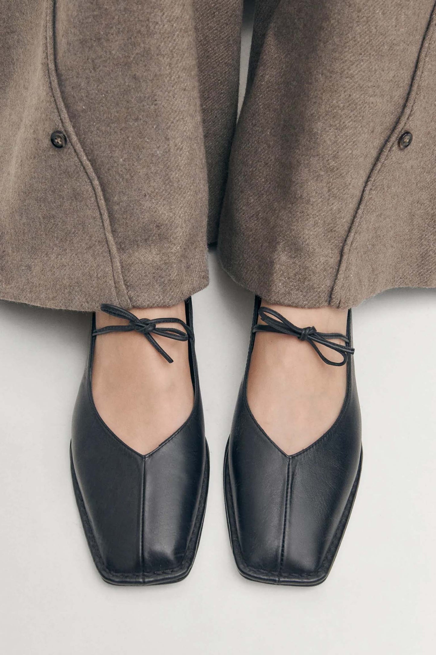 A pair of black leather ballet flats with V-cut vamps and self-tie bows, worn with brown trousers.
