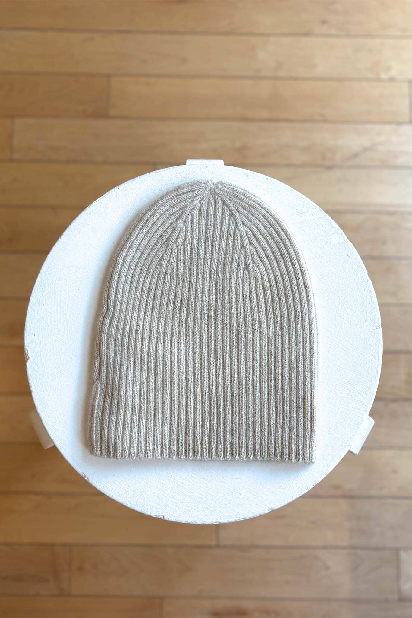 Beige knit beanie on a white cylindrical stand with a wooden floor background