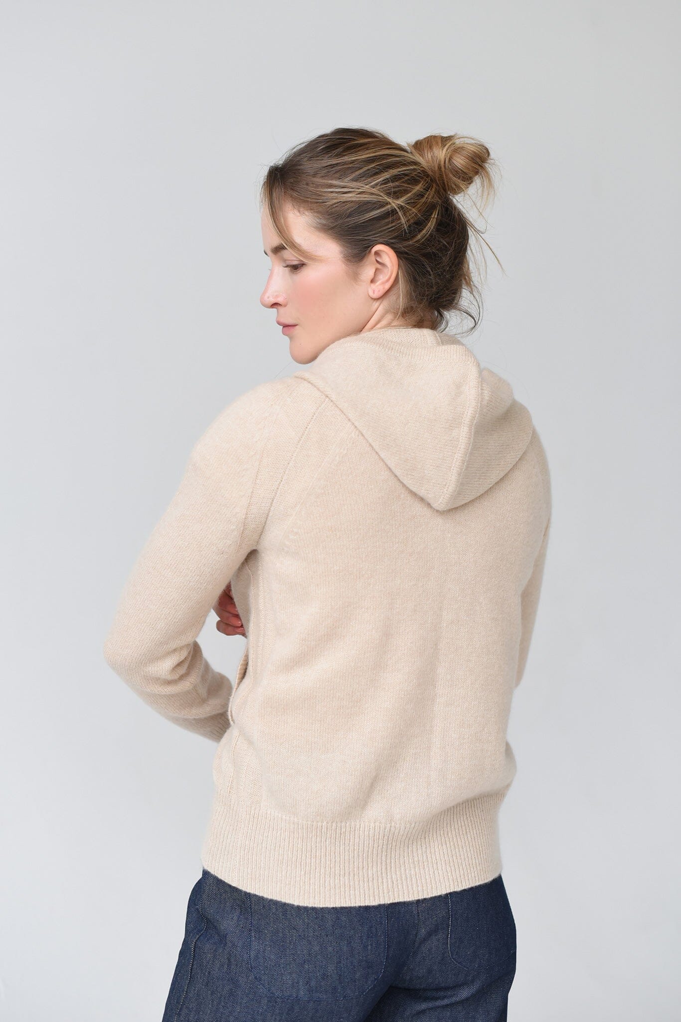 The back of a woman wearing a beige cashmere hoodie with mother of pearl buttons and ribbed trims, paired with a white top and blue jeans.