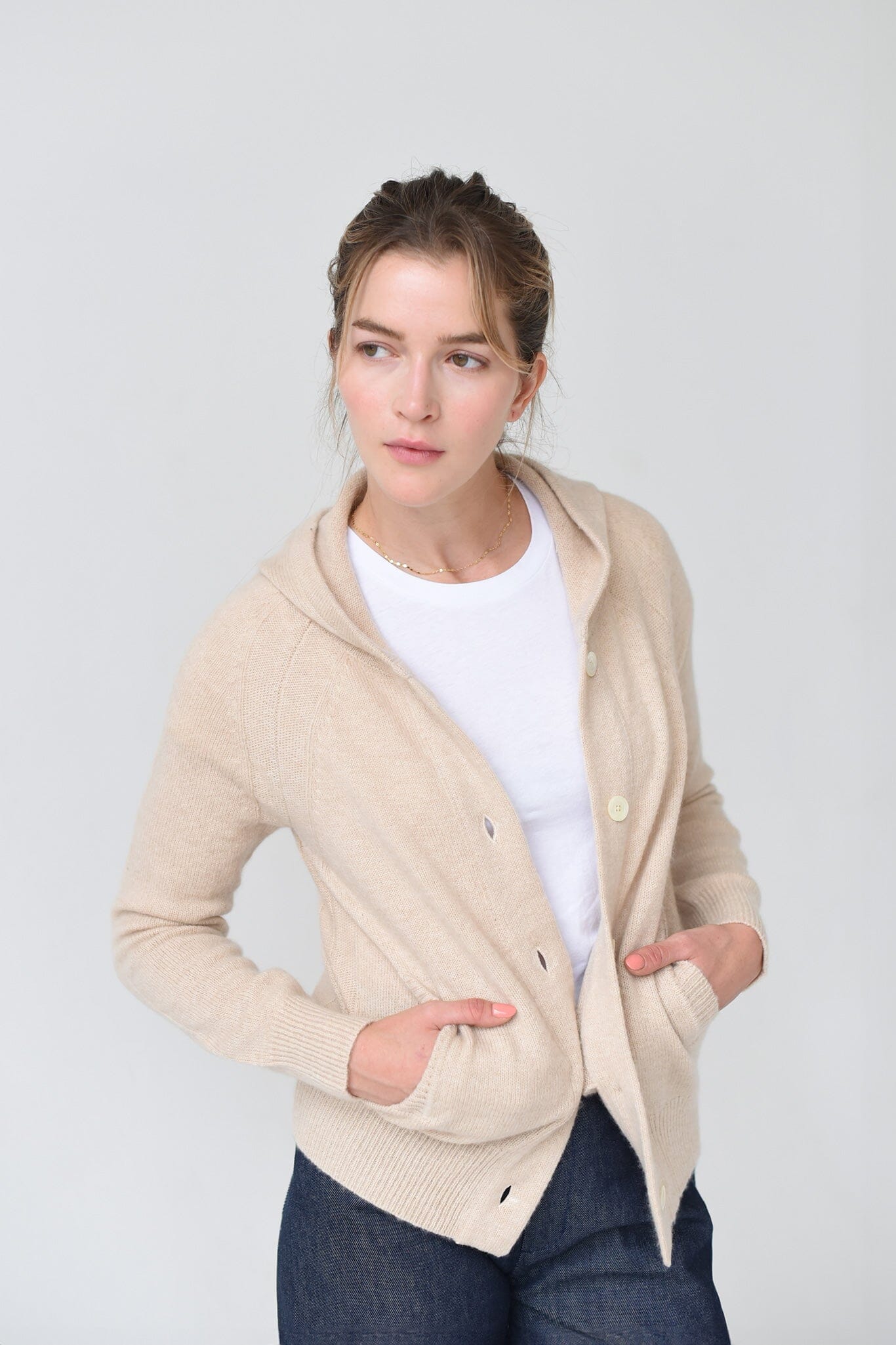 A woman wearing a beige cashmere hoodie with mother of pearl buttons and ribbed trims, paired with a white top and blue jeans.