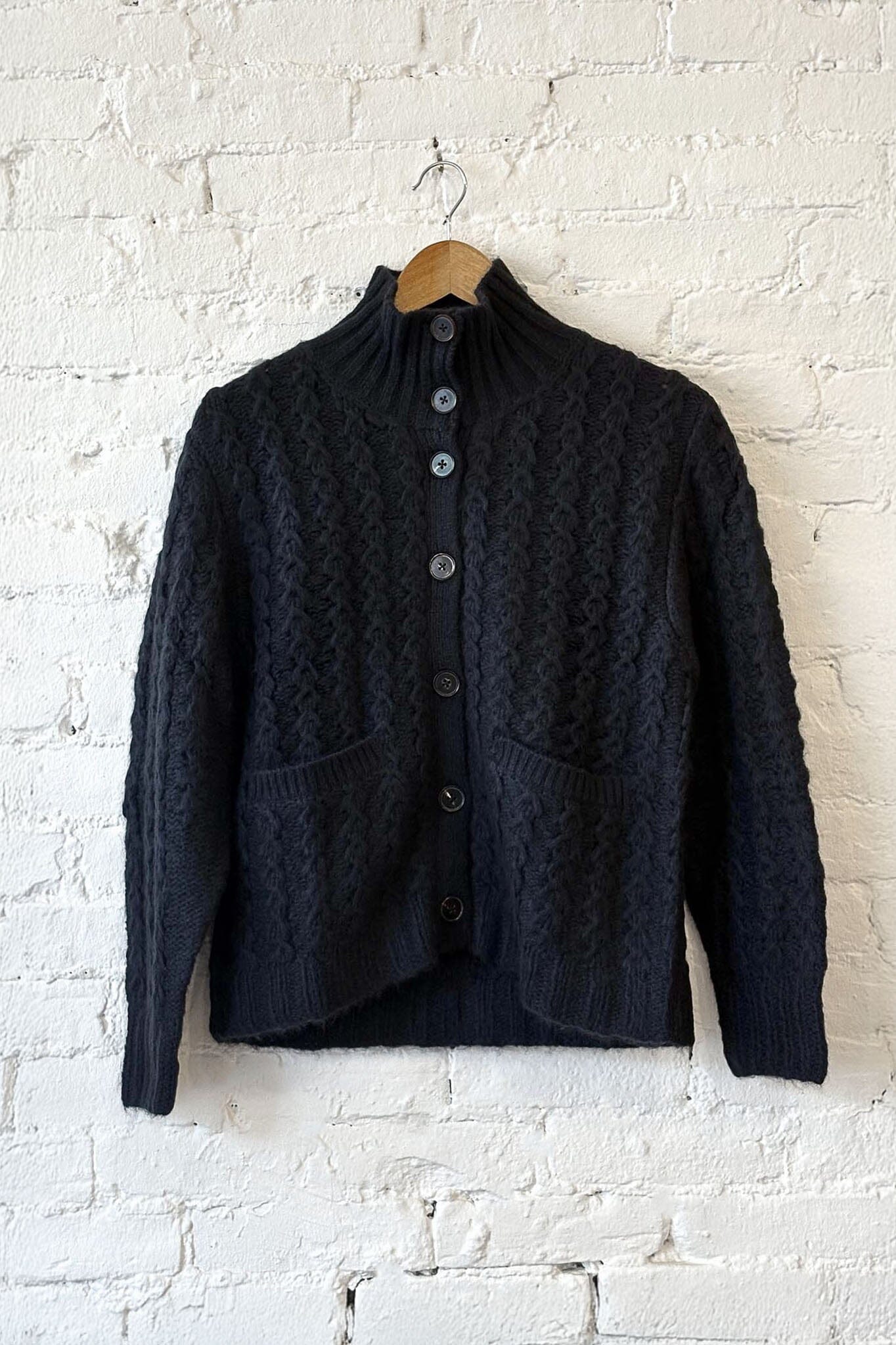 Black knitted cardigan on a wooden hanger against a white brick wall.