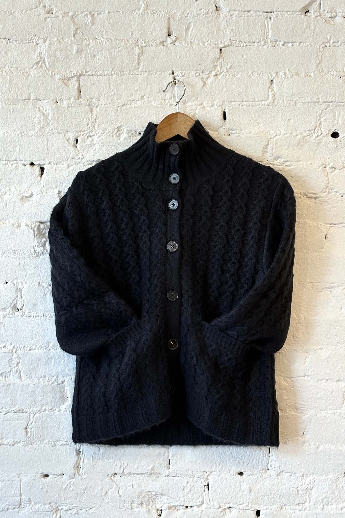 Black knit button down sweater on a wooden hanger against a white brick wall.