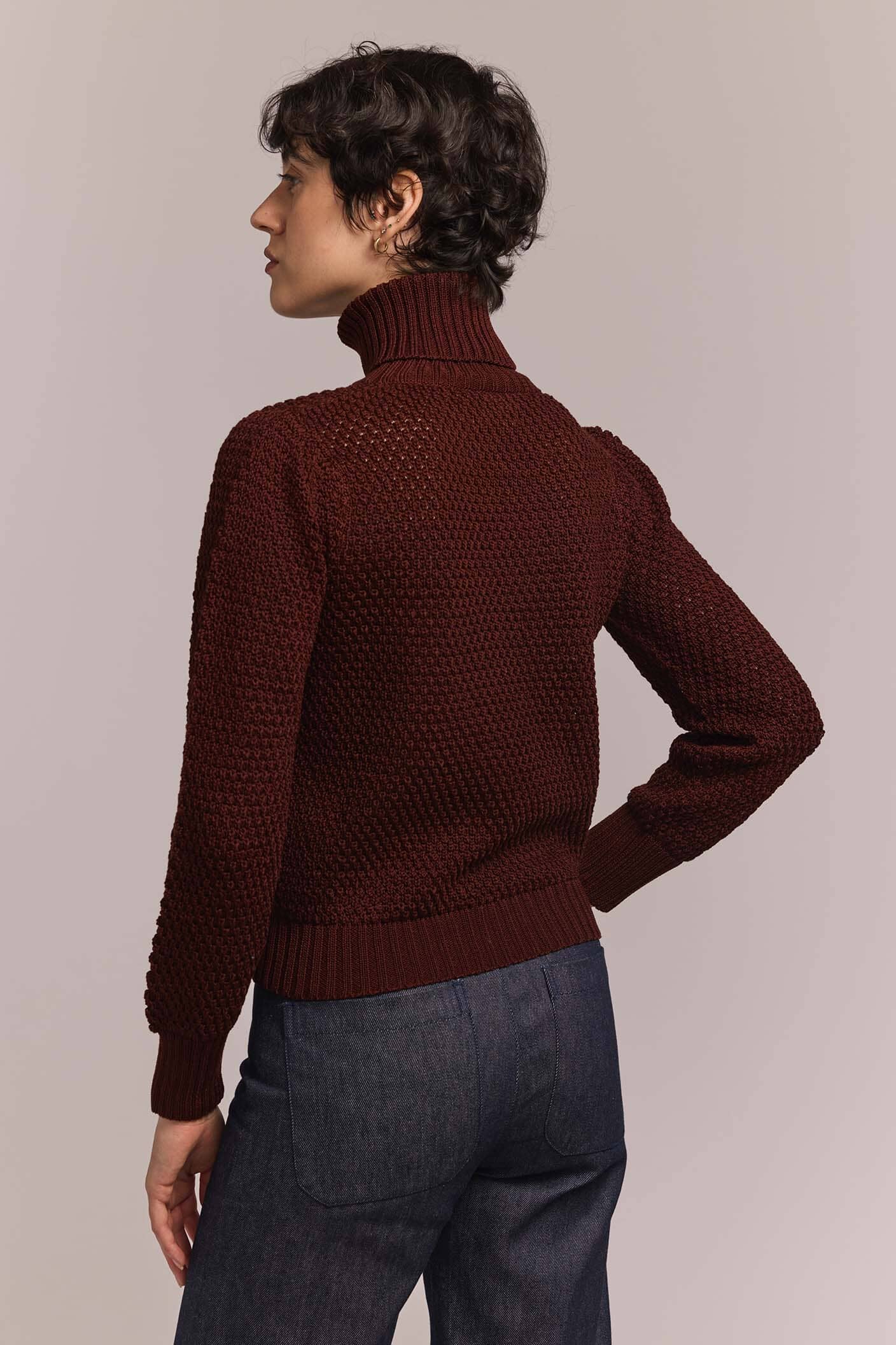 Person wearing a burgundy knitted sweater and dark jeans against a plain background