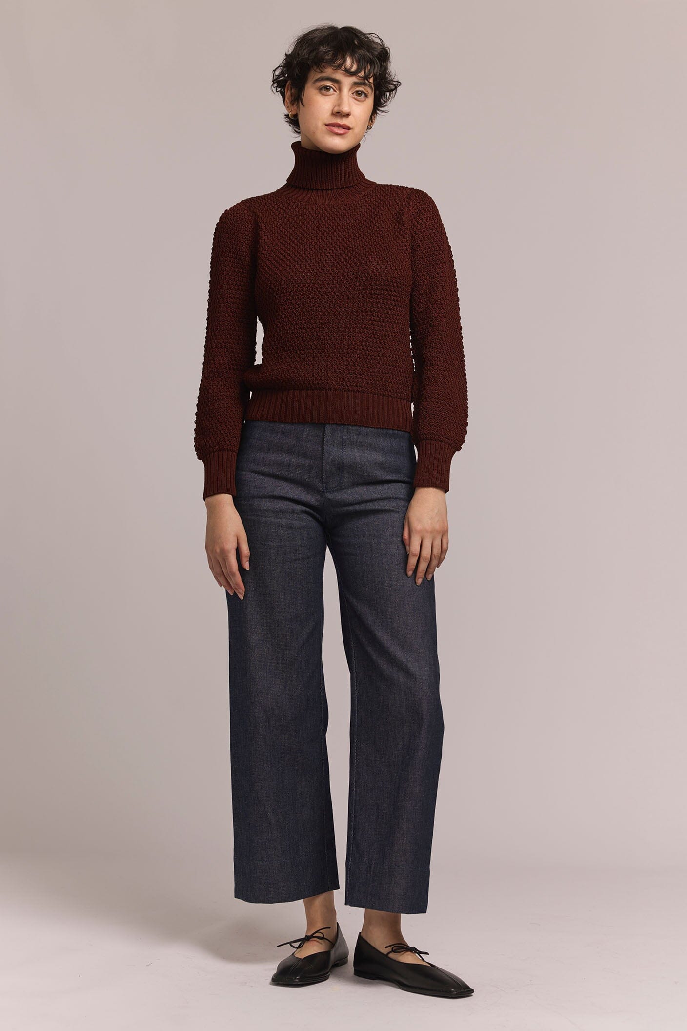 Person wearing a burgundy turtleneck sweater and gray pants on a plain background