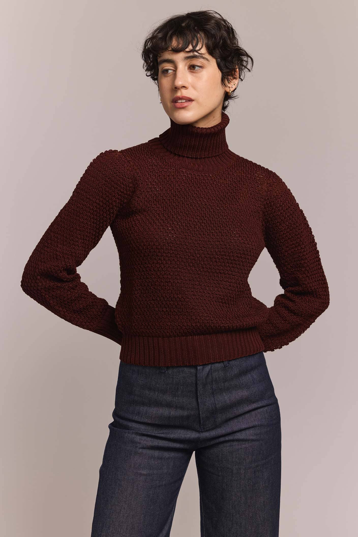 Person wearing a burgundy turtleneck sweater and dark jeans against a plain background