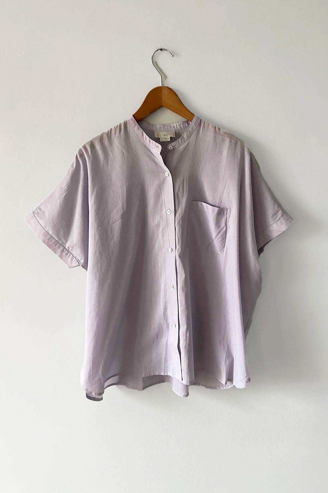 A lavender colored short sleeve blouse with a button-up front displayed on a hanger against a white background.
