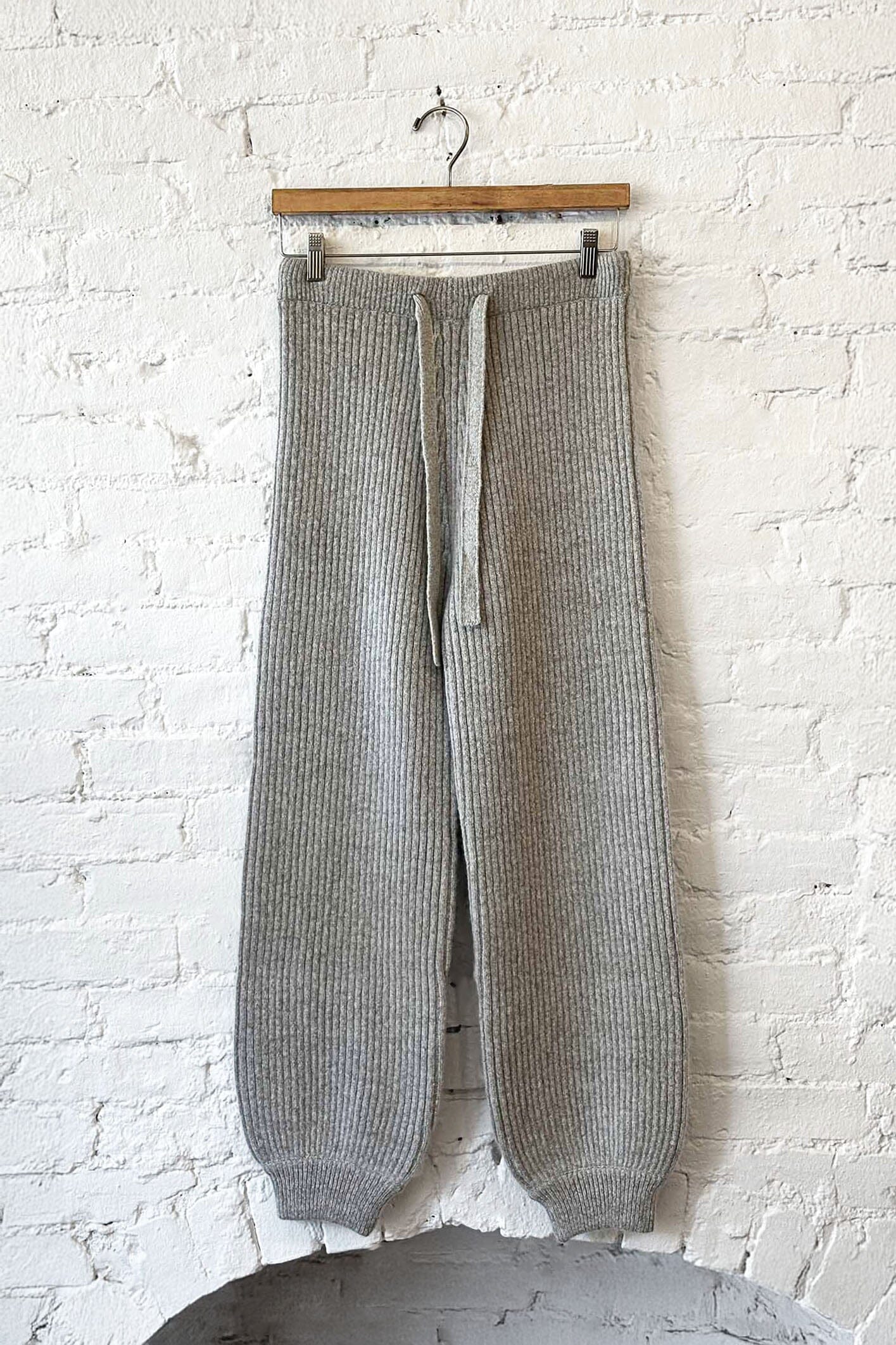 Gray knit pants hanging on a wooden hanger against a white brick wall.