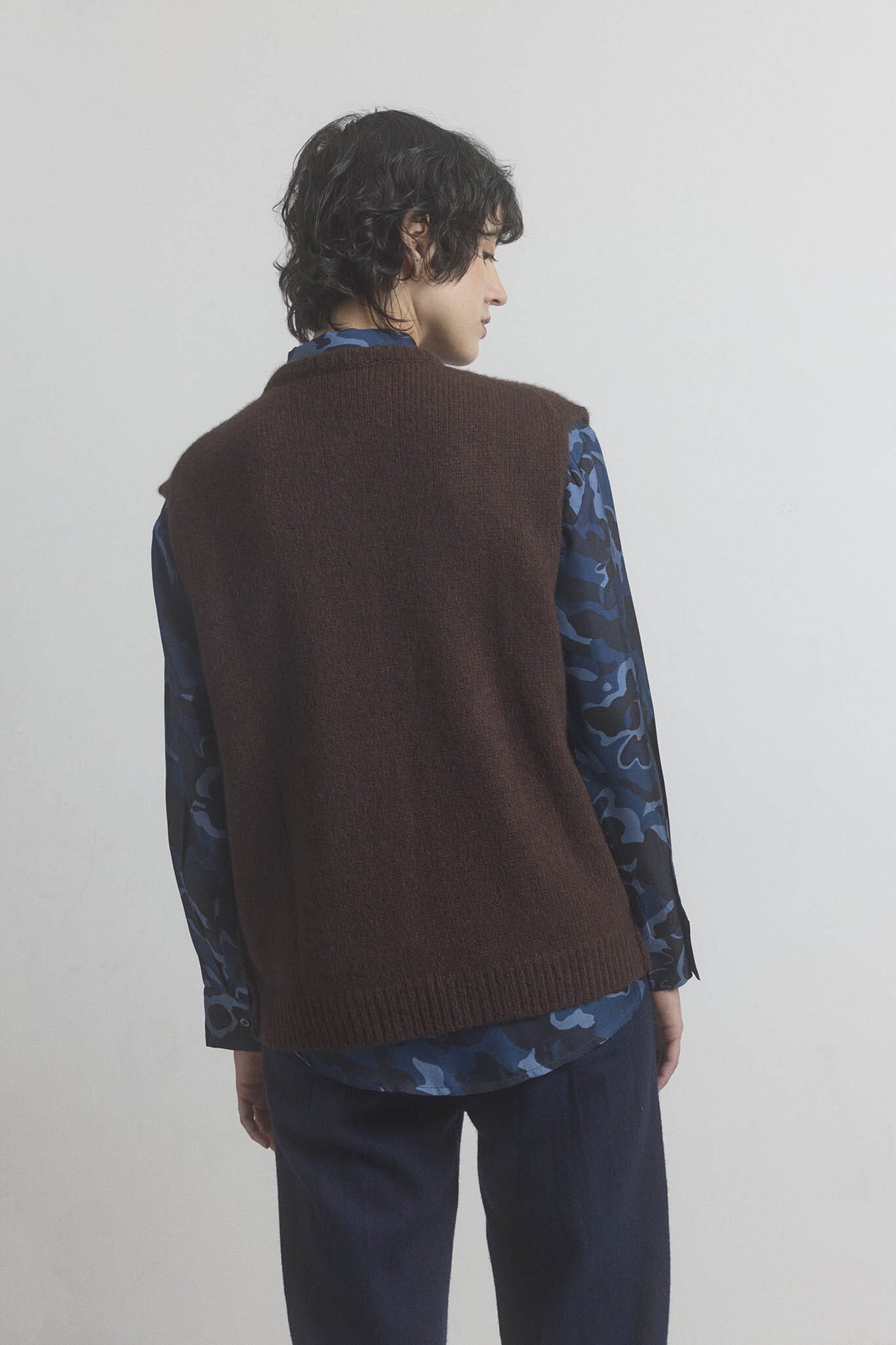 Woman wearing a brown merino vest over a patterned shirt on a plain background