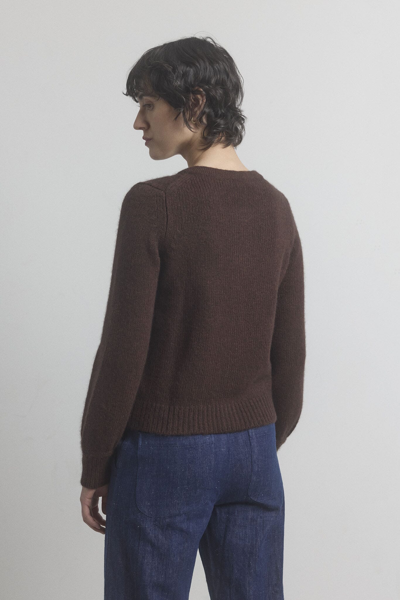 Woman facing the back wearing a brown cashmere sweater and blue pants against a plain background