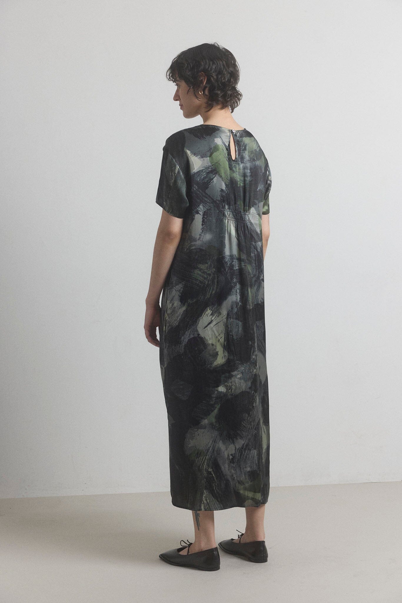 Woman facing the back wearing a long, dark green silk dress with a abstract pattern on a plain background