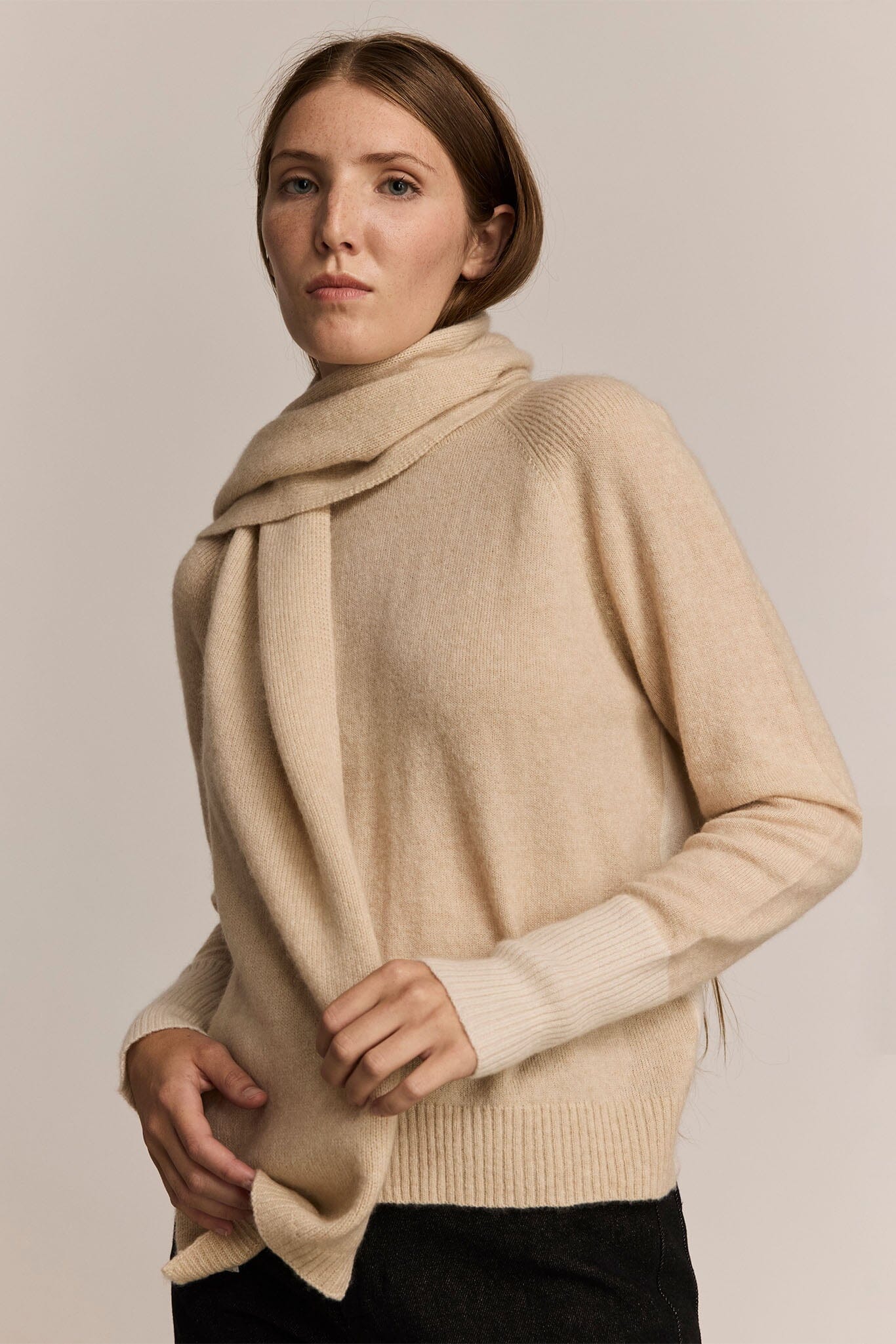 Woman wearing a long beige knit sweater and scarf against a plain background
