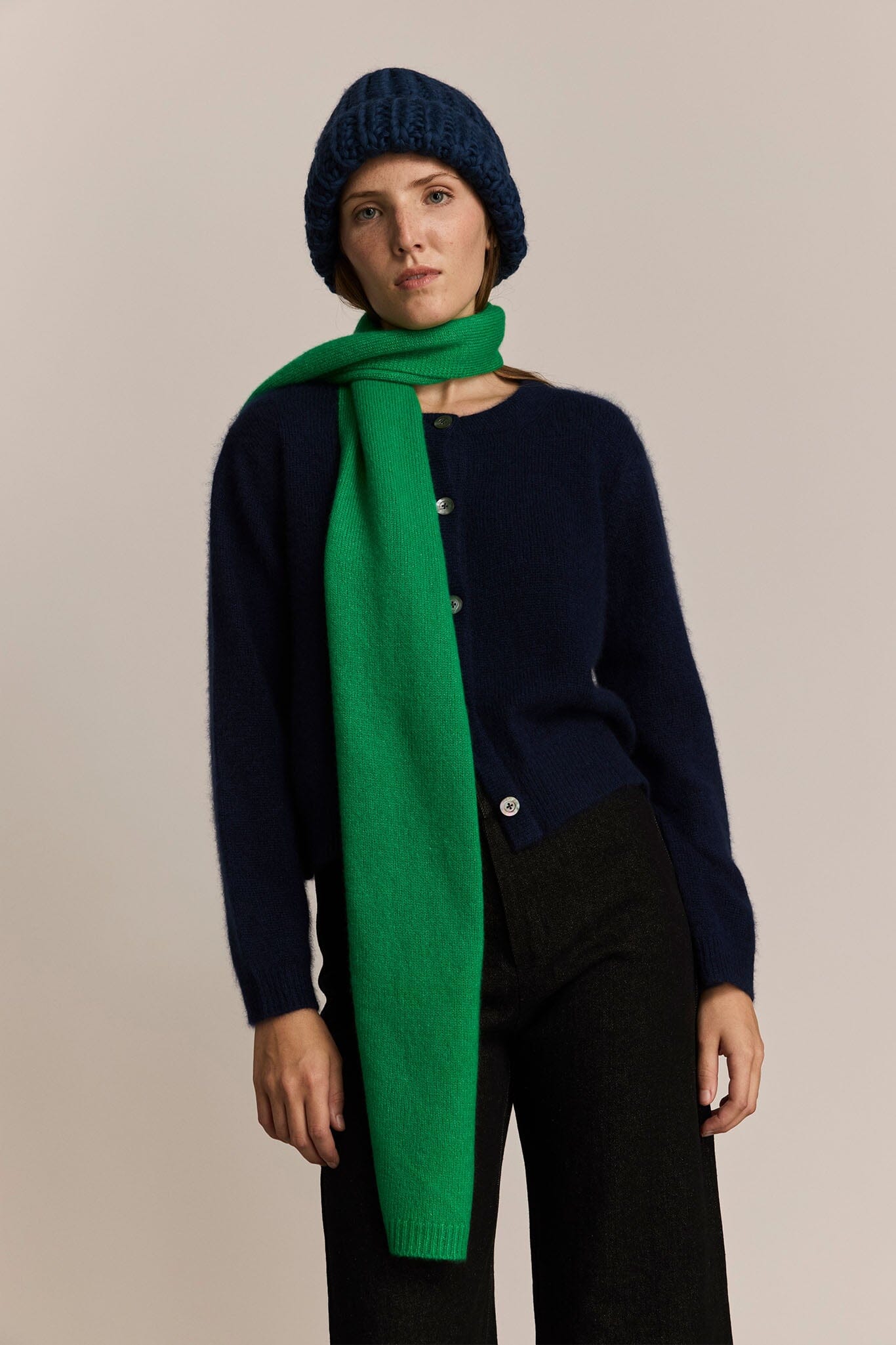 Woman wearing a navy sweater, black pants, and a long green scarf with a beige background