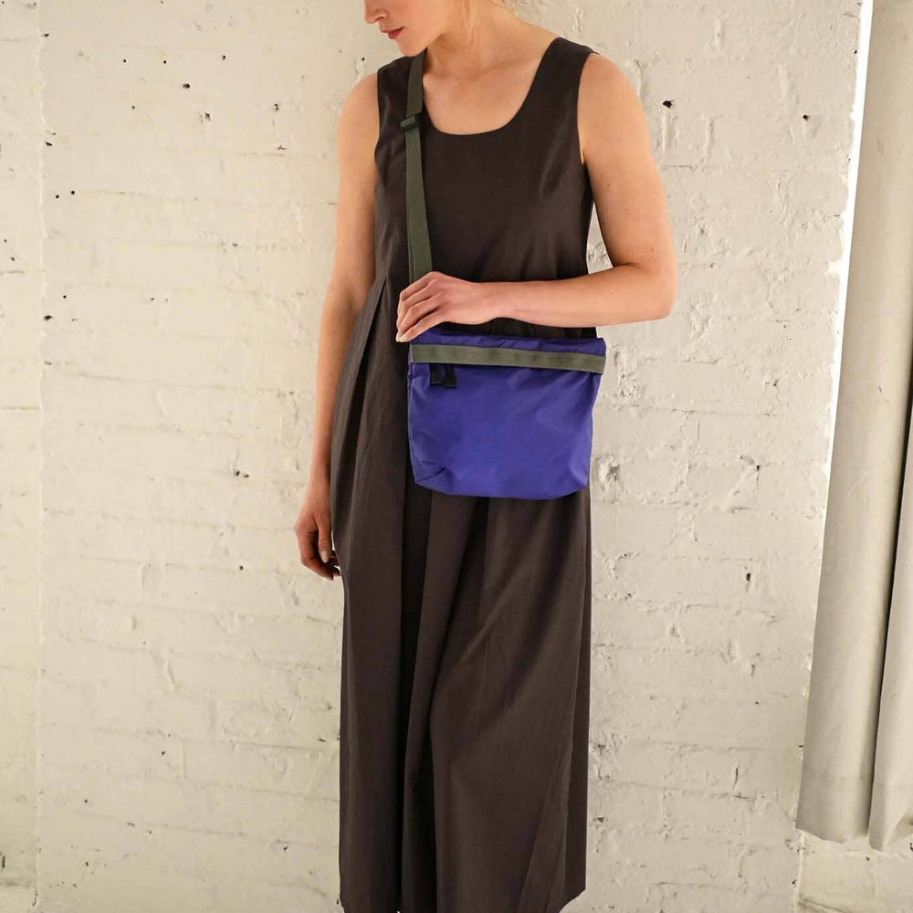 Woman wearing a long brown dress with a purple crossbody bag over her shoulder against a white brick wall.