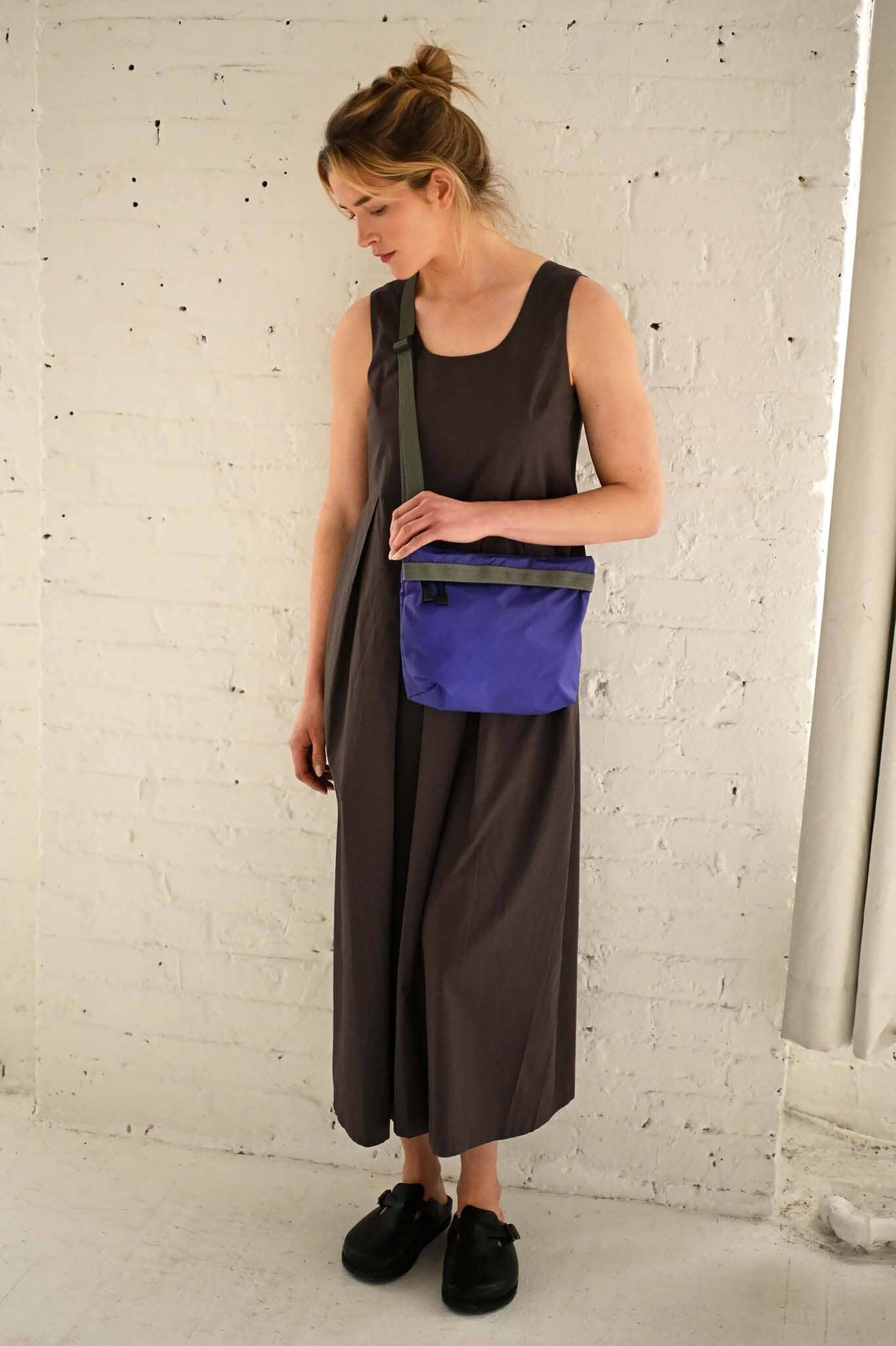 Woman wearing a long brown dress with a purple crossbody bag over her shoulder against a white brick wall.
