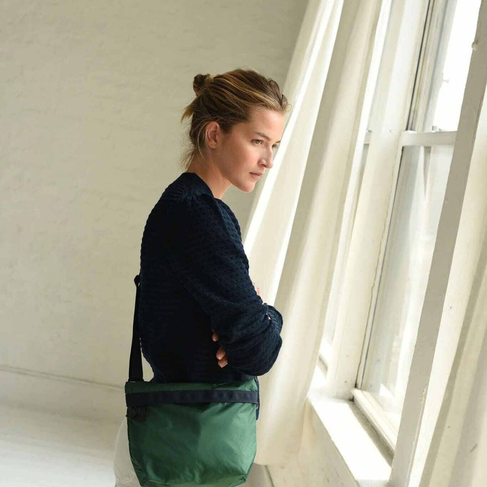 A woman with a blue sweater stands with a forest green nylon crossbody bag against a cream background