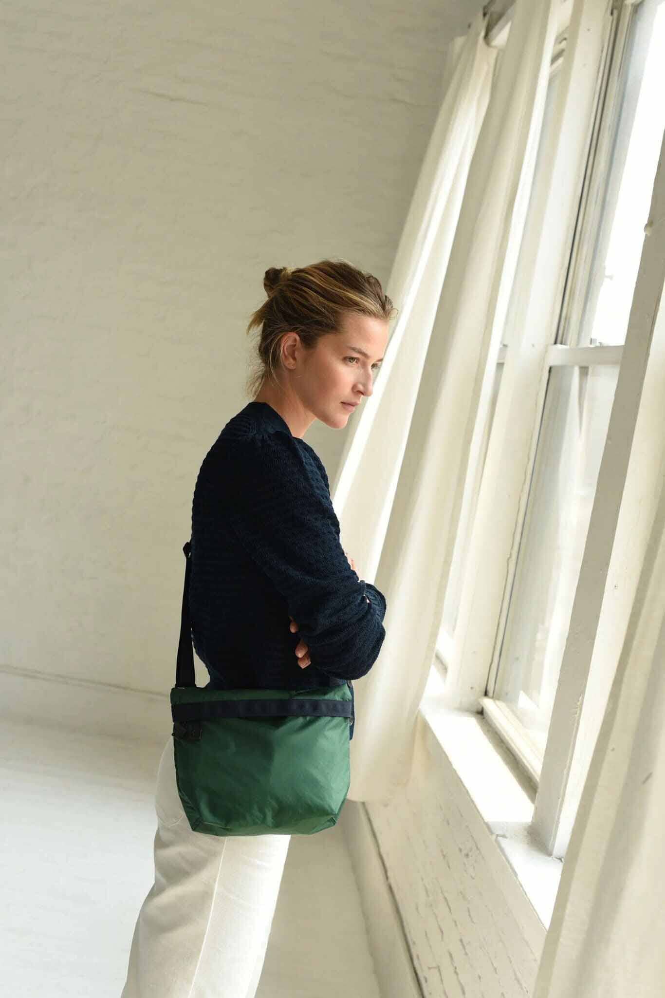 A woman with a blue sweater stands with a forest green nylon crossbody bag against a cream background
