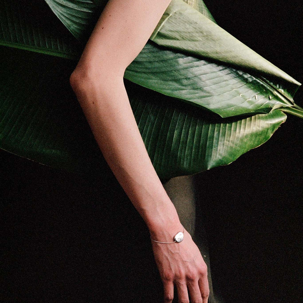 
                  
                    Editorial image of a woman wearing a silver pendent bracelet with a large green leaf on a dark background
                  
                