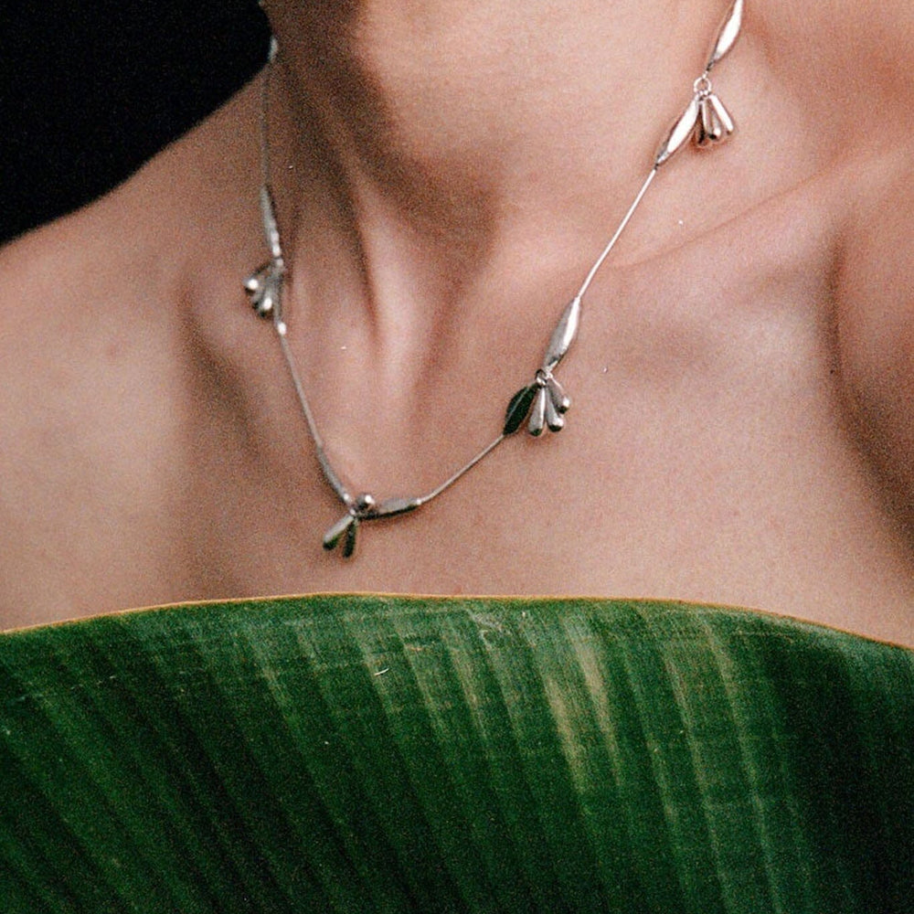 
                  
                    Silver necklace with floral leaf designs worn by a person against a dark background
                  
                