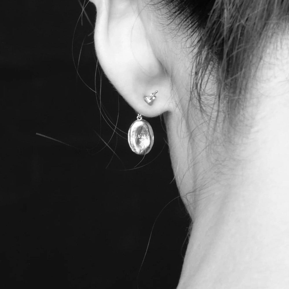 
                  
                    Close-up of an ear wearing a silver drop stud earring against a dark background
                  
                