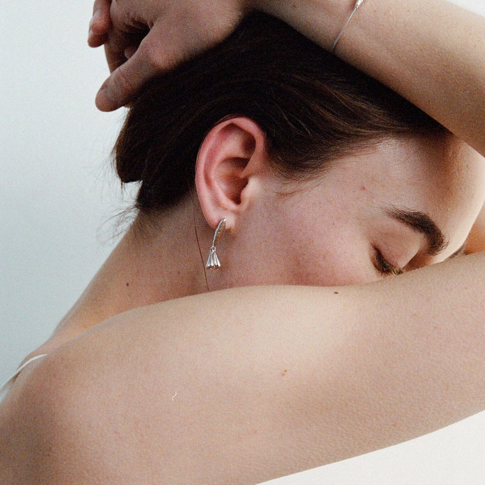 Woman with one arm around her head and the other arm draped over her shoulder, wearing sterling silver floral stud earrings against a light background