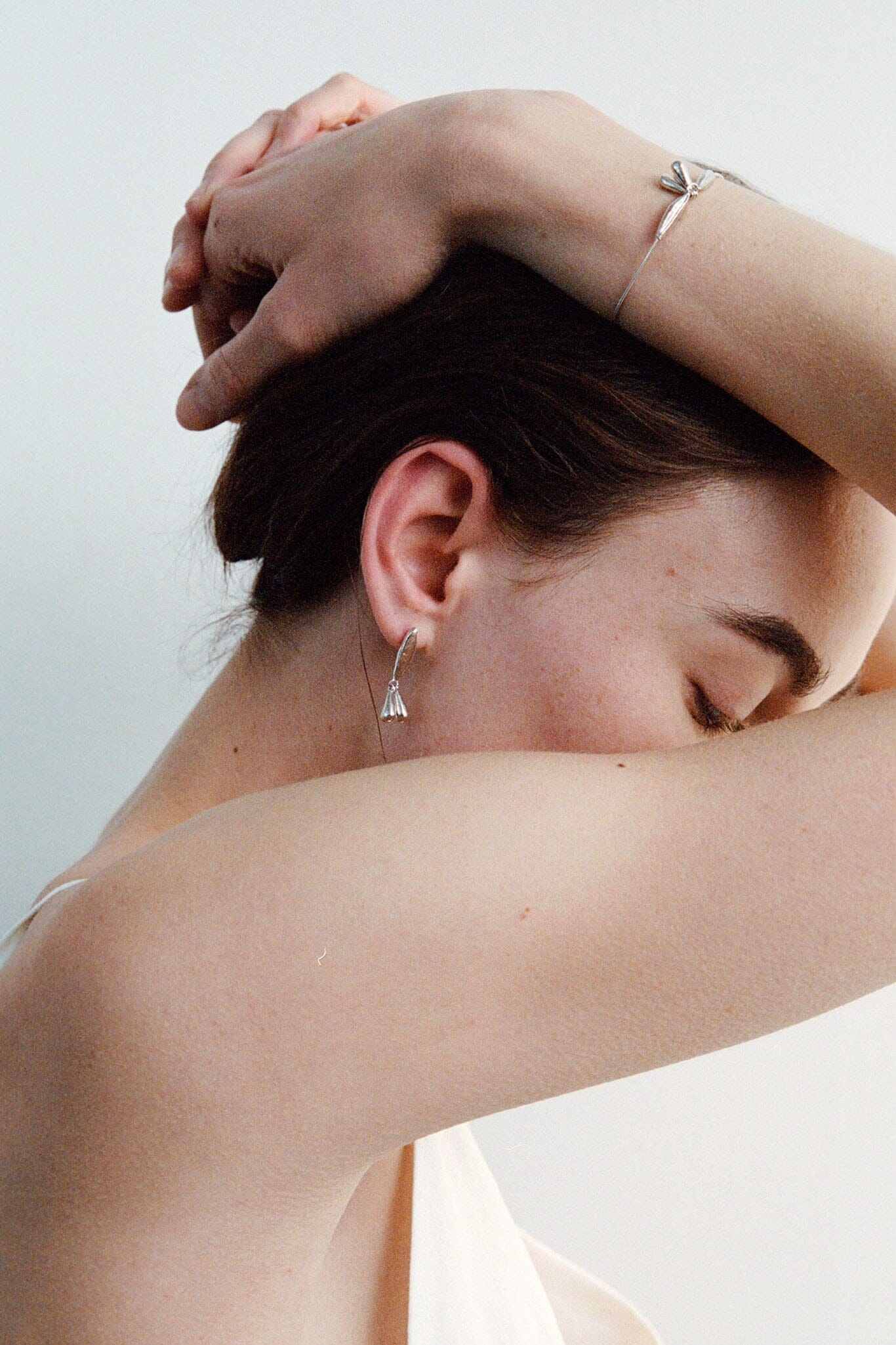 Woman with one arm around her head and the other arm draped over her shoulder, wearing sterling silver floral stud earrings against a light background