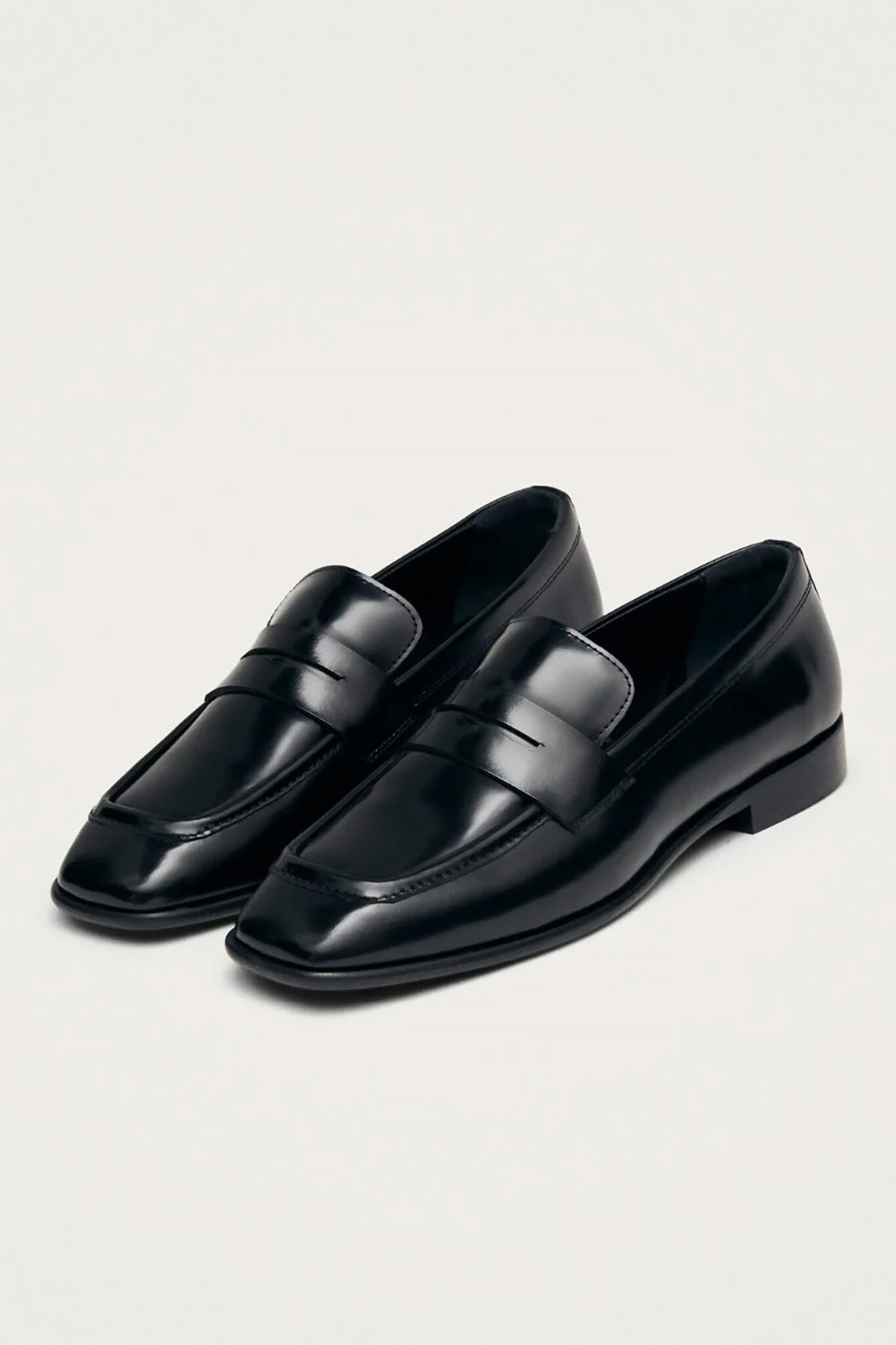 A pair of black leather penny loafers with a glossy finish, shown from a high angle on a light off-white background.