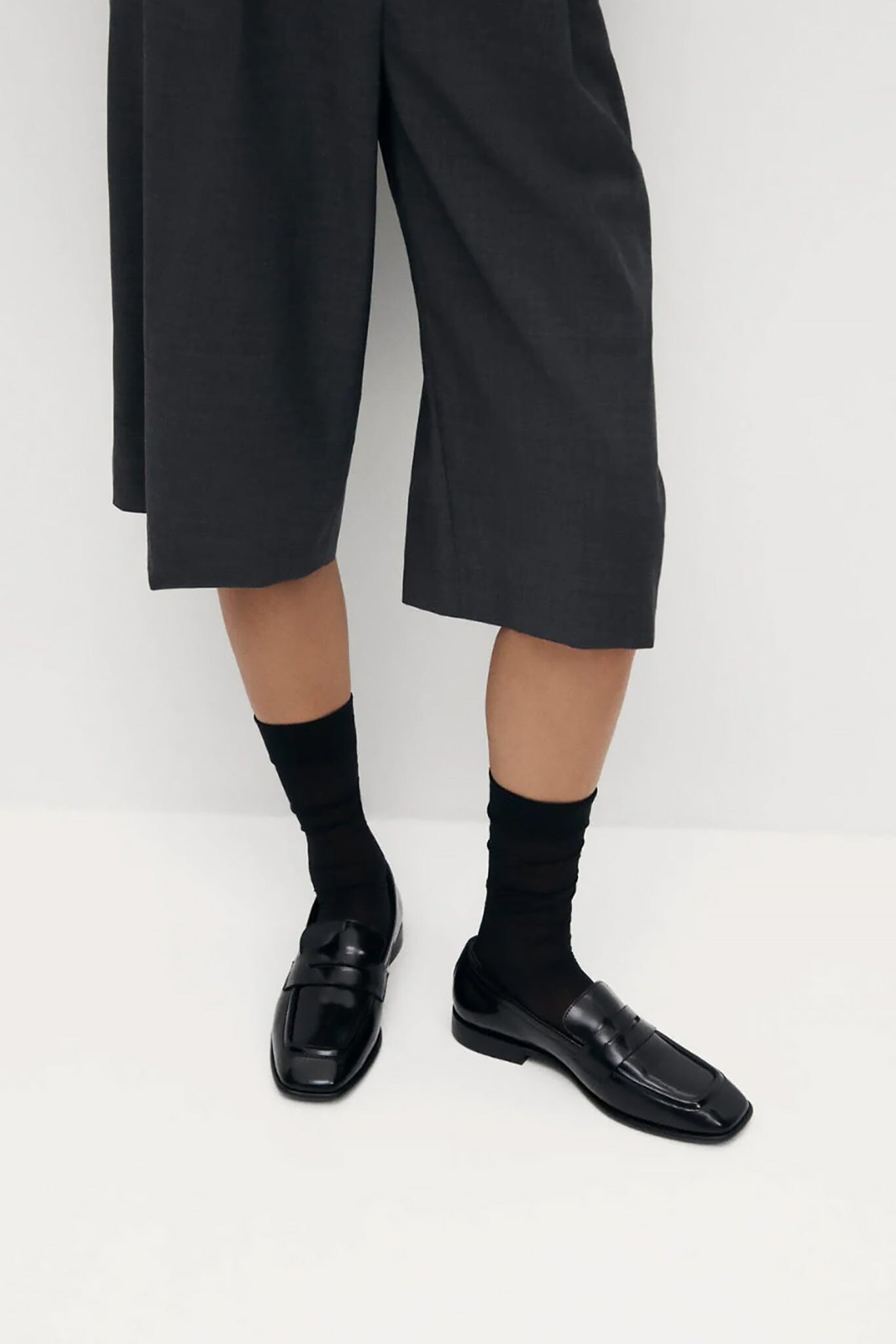 A person wearing black cropped wide-leg trousers, black crew socks, and black leather penny loafers, shown from the waist down against a plain light background.
