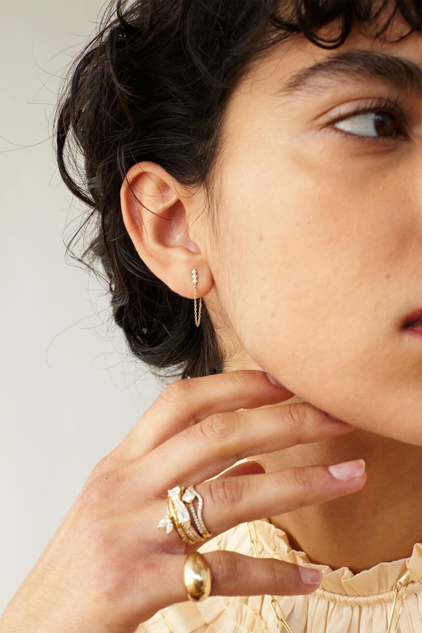 Close-up of a person wearing gold hoop earrings and rings on a neutral background