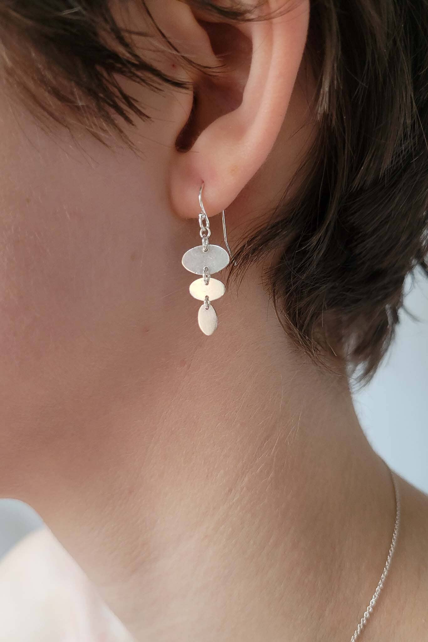 Close-up of an ear wearing a silver earring with dangling oval geometric shapes.