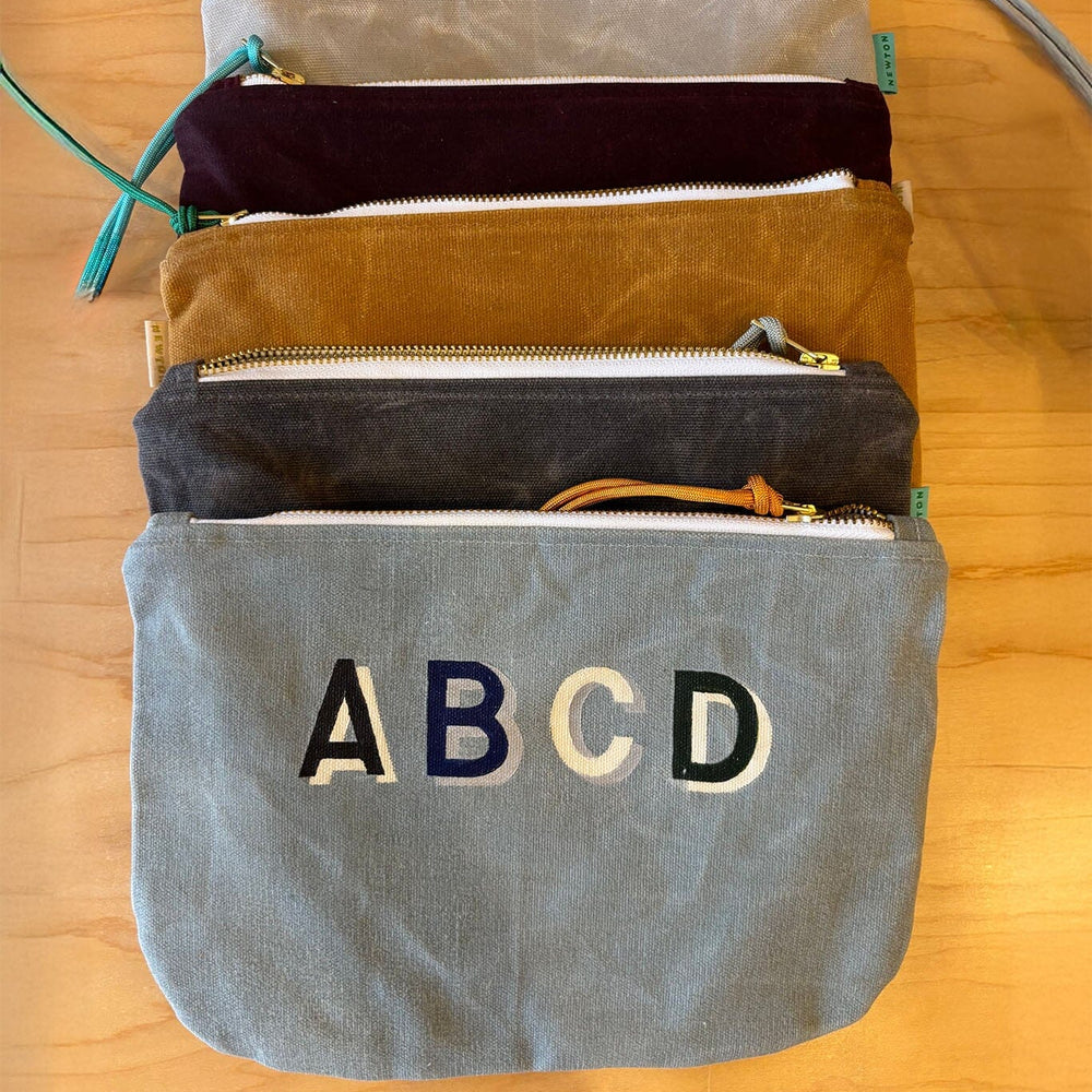 
                  
                    Set of four pouches with zippers on a wooden surface, with 'ABCD' embroidered on the largest pouch.
                  
                