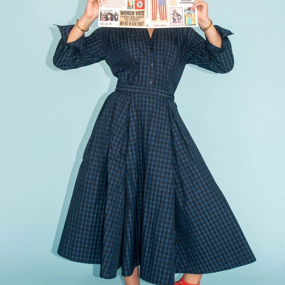 
                  
                    Person wearing a dark blue checkered dress with red socks holding a newspaper against a light blue background
                  
                