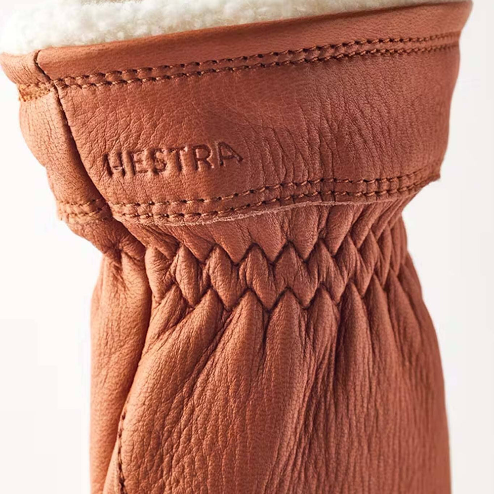 
                  
                    Close-up of a brown leather glove with white fur lining, featuring the Hestra brand logo.
                  
                
