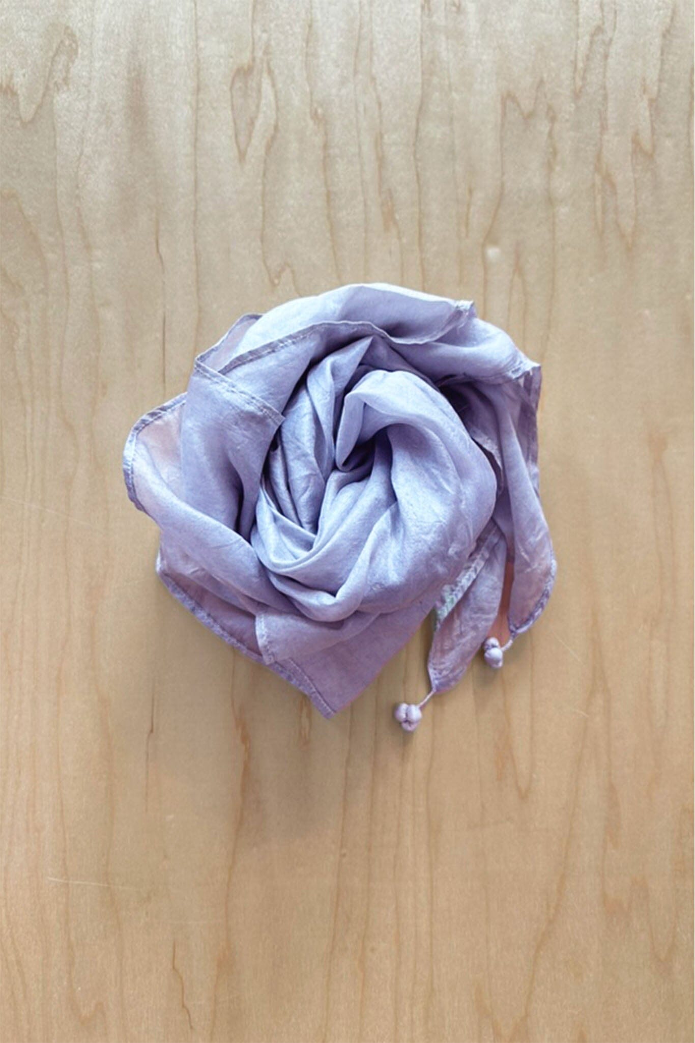 Folded light purple silk scarf with tassel accents on a wooden surface