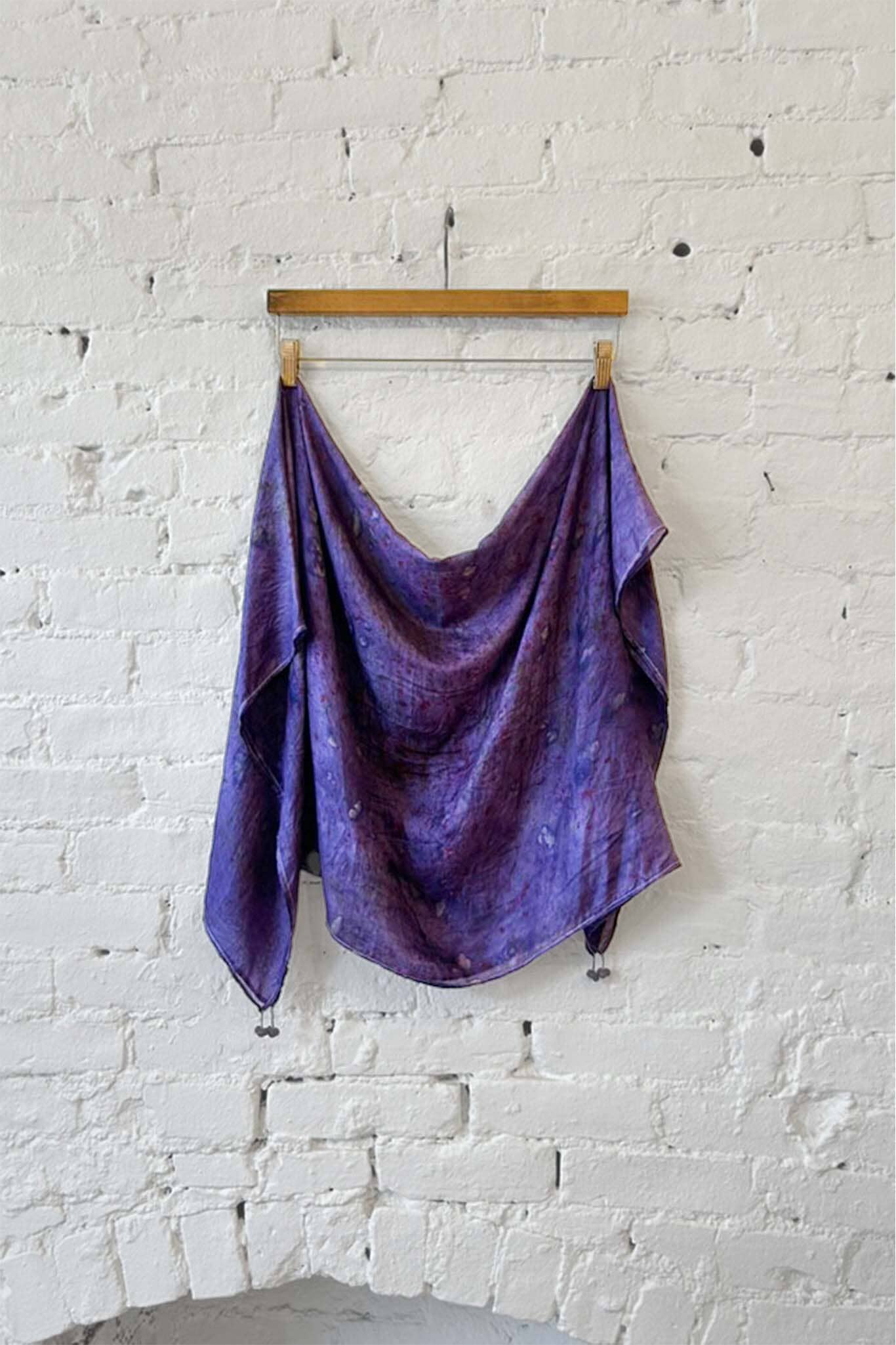 Purple silk scarf draped over a wooden hanger against a white brick wall.