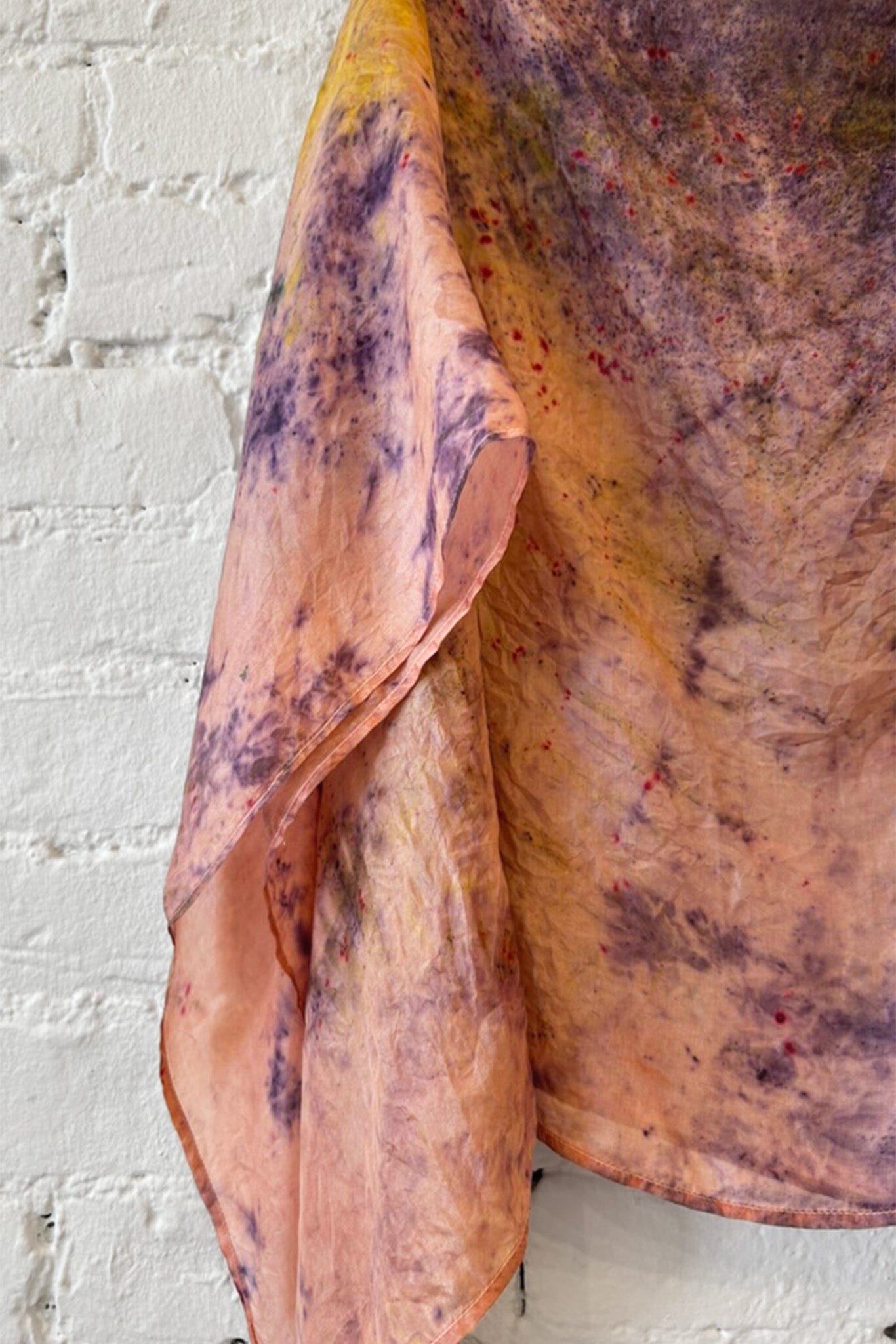 Tie dye silk scarf with earthy tones draped over a surface against a white brick wall.