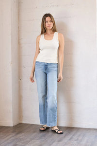 Woman wearing a white tank top and light blue jeans standing against a plain wall.
