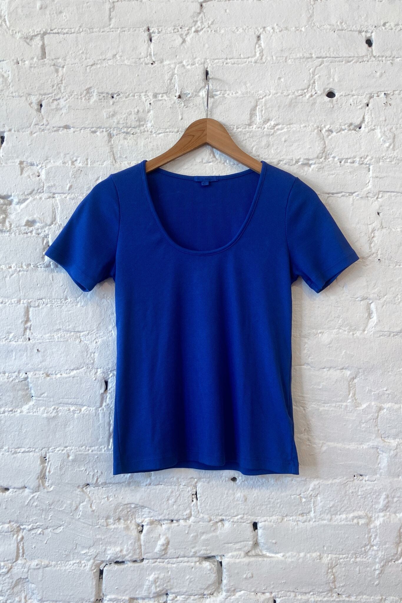 A royal blue short-sleeve t-shirt hanging on a wooden hanger against a white painted brick wall.