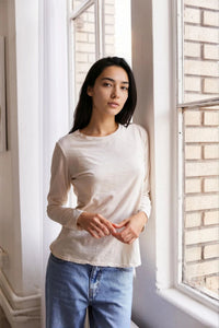 Woman wearing a cream colored long-sleeve shirt and blue jeans standing in a room with a window and brick wall.